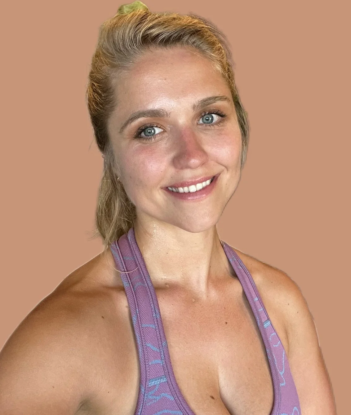 A smiling woman with light blonde hair and blue eyes wearing a purple sports top against a beige background.