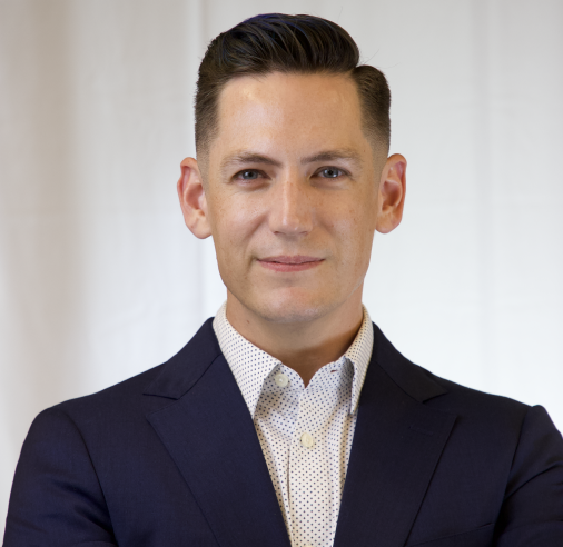 Man in business suit with white shirt looking at camera, standing against a plain background.