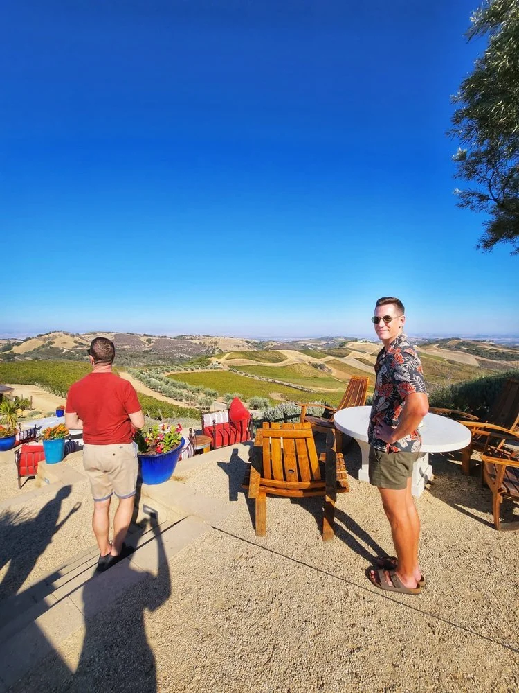 Two men standing on a patio with outdoor furniture, overlooking rolling hills and farmland under a clear blue sky.