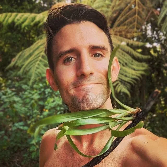A man outdoors holding a plant with long green leaves close to his shoulder, with dense greenery in the background.