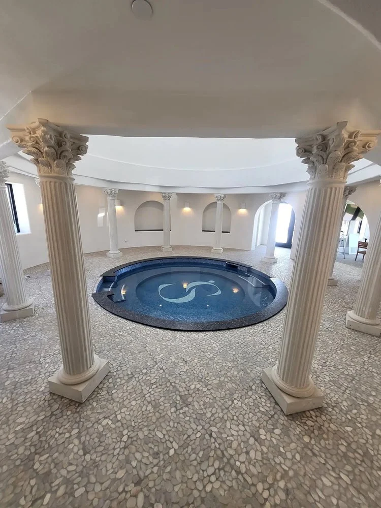 Indoor spa with circular hot tub, surrounded by Roman-style columns and stone mosaic flooring.