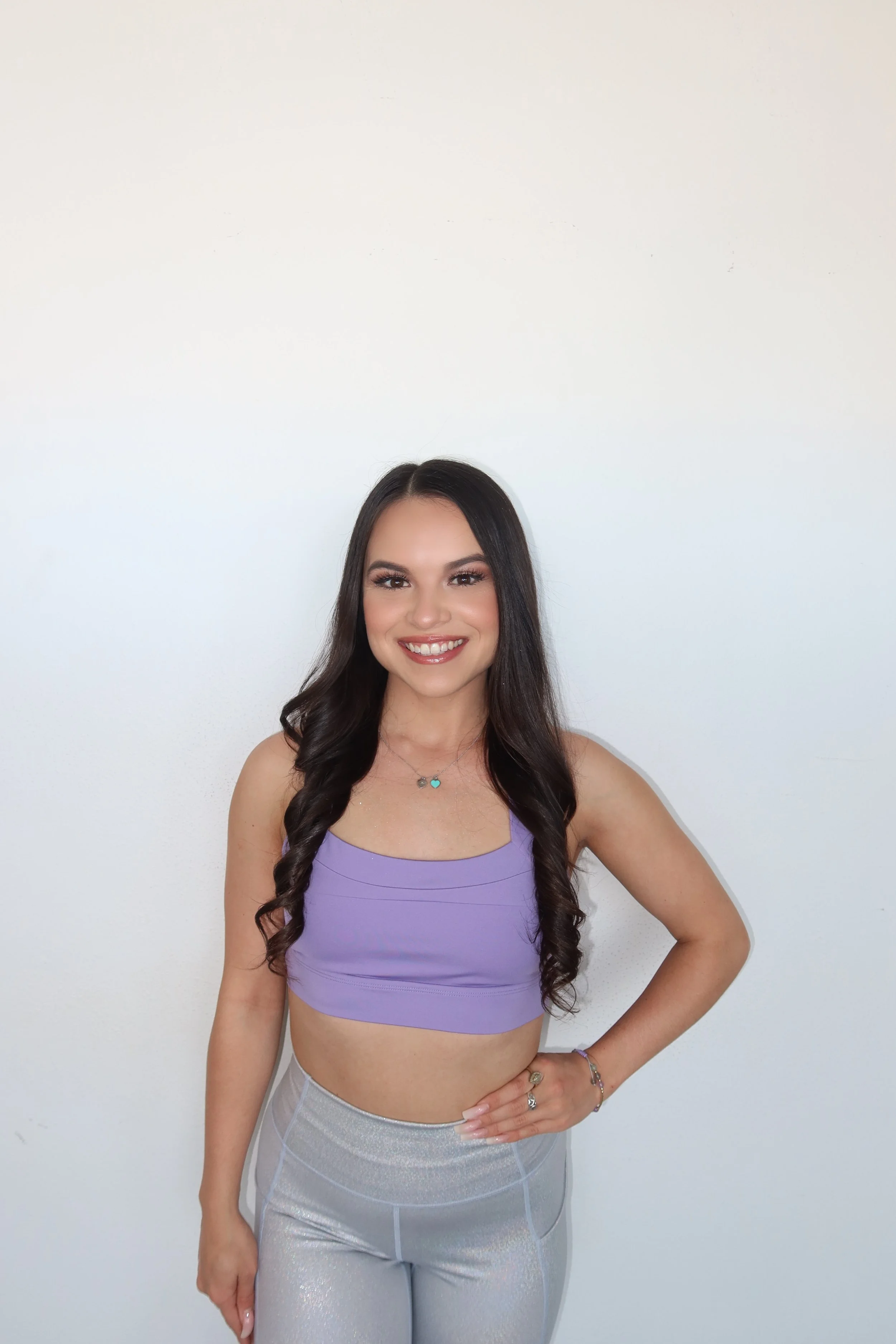 A young woman with long dark hair, wearing a purple crop top and shiny silver leggings, posing against a plain white background, smiling with one hand on her hip.