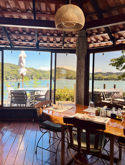 Restaurant interior with wooden tables set for dining, overlooking a scenic lake view through large windows. Outdoor seating with umbrellas is visible. The setting is bright with natural light.