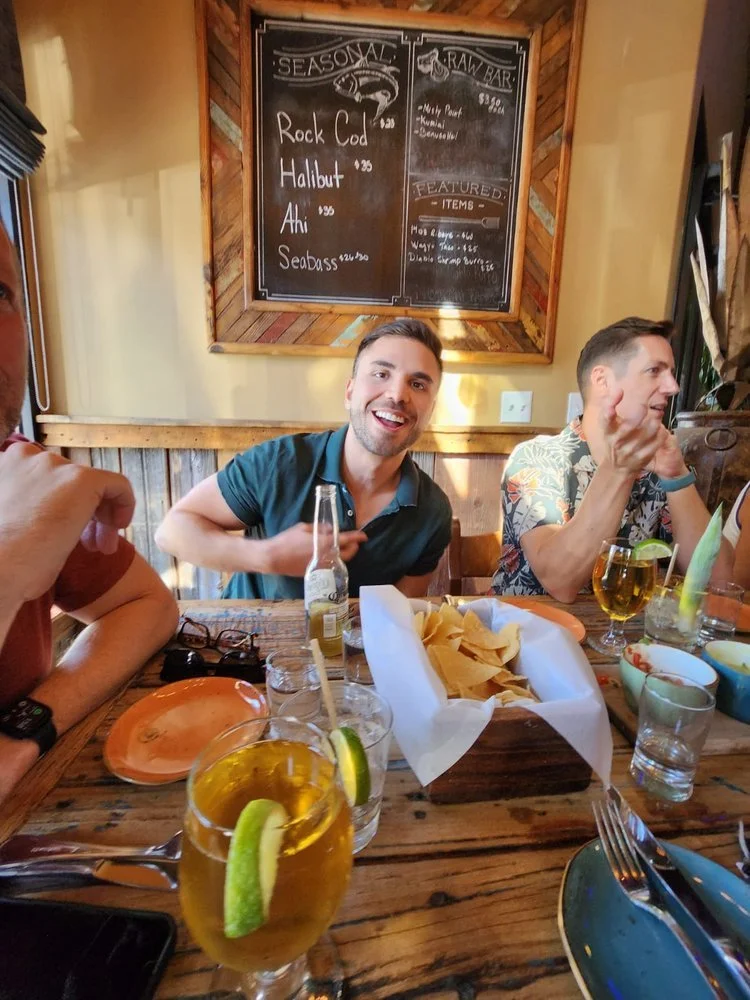 People dining at a restaurant, with drinks and chips on the table, and a menu board in the background.