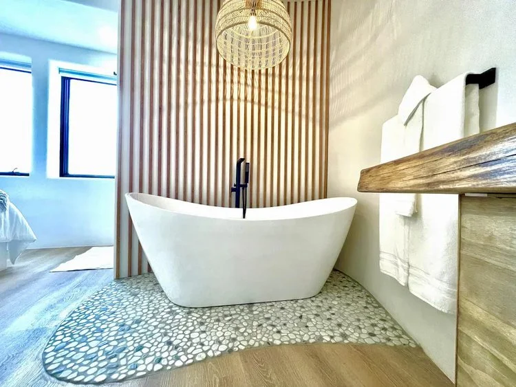 Modern bathroom with freestanding white bathtub, wooden slat wall, pebble stone floor accents, towel rack, and ceiling pendant light.