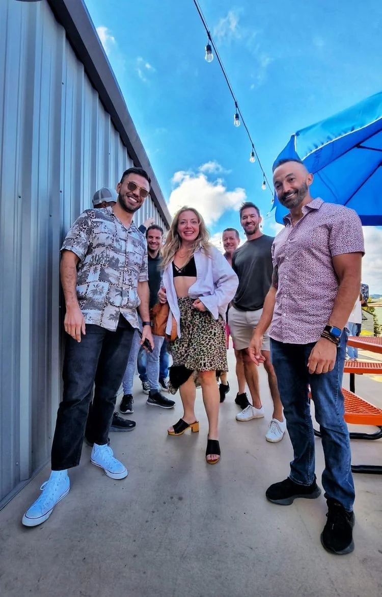 Group of people smiling outdoors on a sunny day, standing on a patio. They are casually dressed, with a metal wall on one side and blue sky with clouds above.