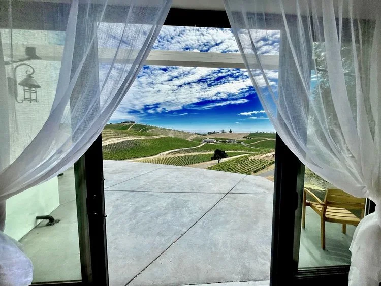 View of vineyard through open patio doors with sheer curtains, showing rolling hills, a clear sky, and outdoor seating.