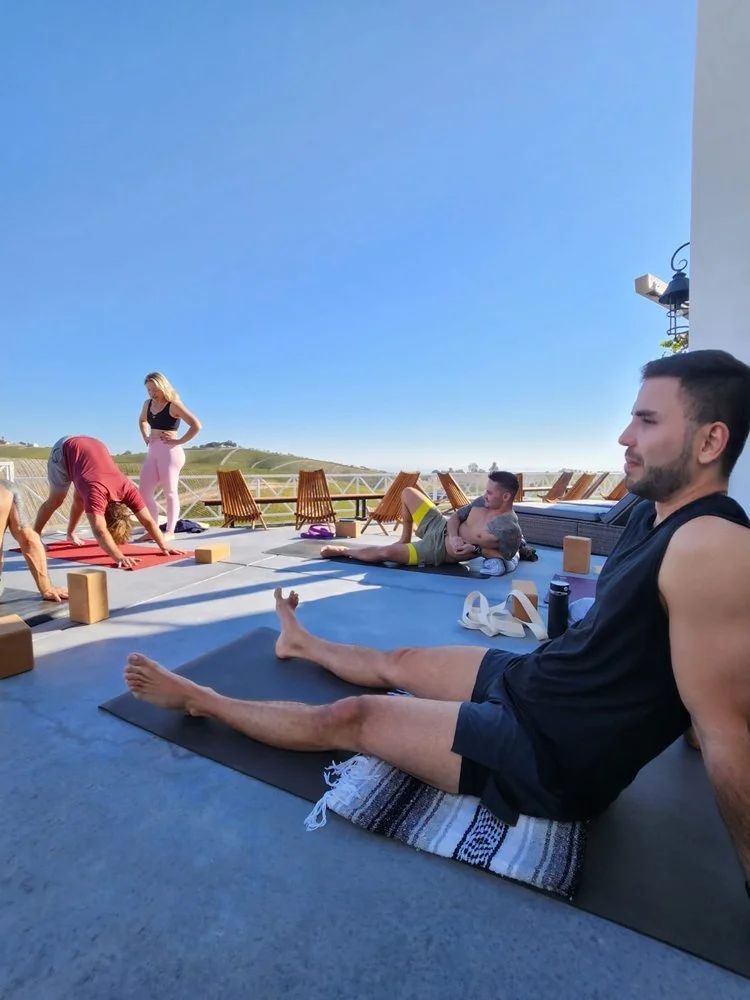 People practicing yoga outdoors on a sunny patio, some performing poses on mats and others sitting. Yoga blocks and water bottles are visible, with a scenic view in the background.