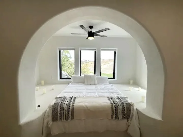 Minimalist bedroom with archway, white bedding, decorative pillows, ceiling fan, and three large windows with a scenic view.