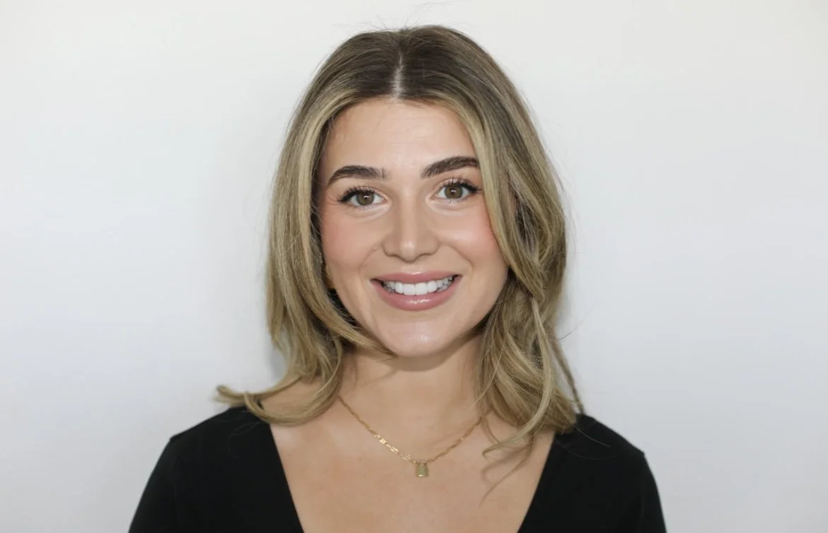 Close-up photo of a smiling young woman with shoulder-length blonde hair, wearing a black top and a gold necklace, against a plain white background.