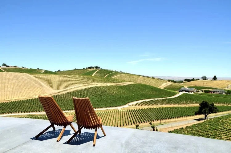 Two wooden chairs on a terrace overlooking a vineyard with rolling hills under a clear blue sky.