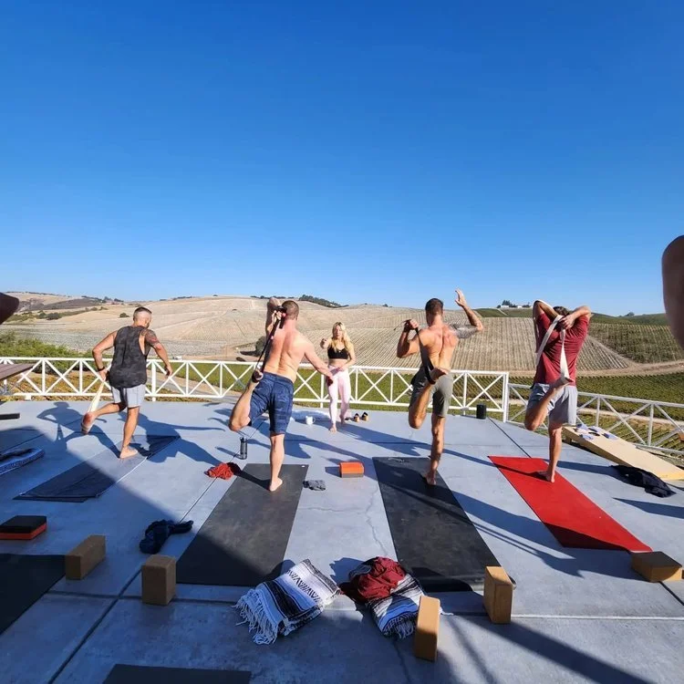 Group of people practicing yoga outdoors on mats in a scenic area with hills in the background.