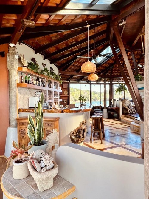 Rustic home interior with wooden beams, a bar area with bottles, potted plants, and wicker pendant lights. Large windows reveal exterior views. Decor includes natural elements like shells and driftwood.
