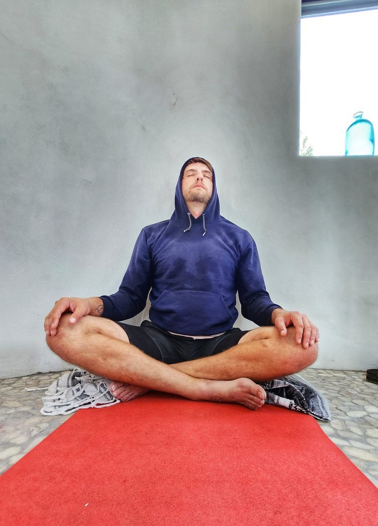 Person meditating in a seated position on a red mat, wearing a blue hoodie, eyes closed, legs crossed.