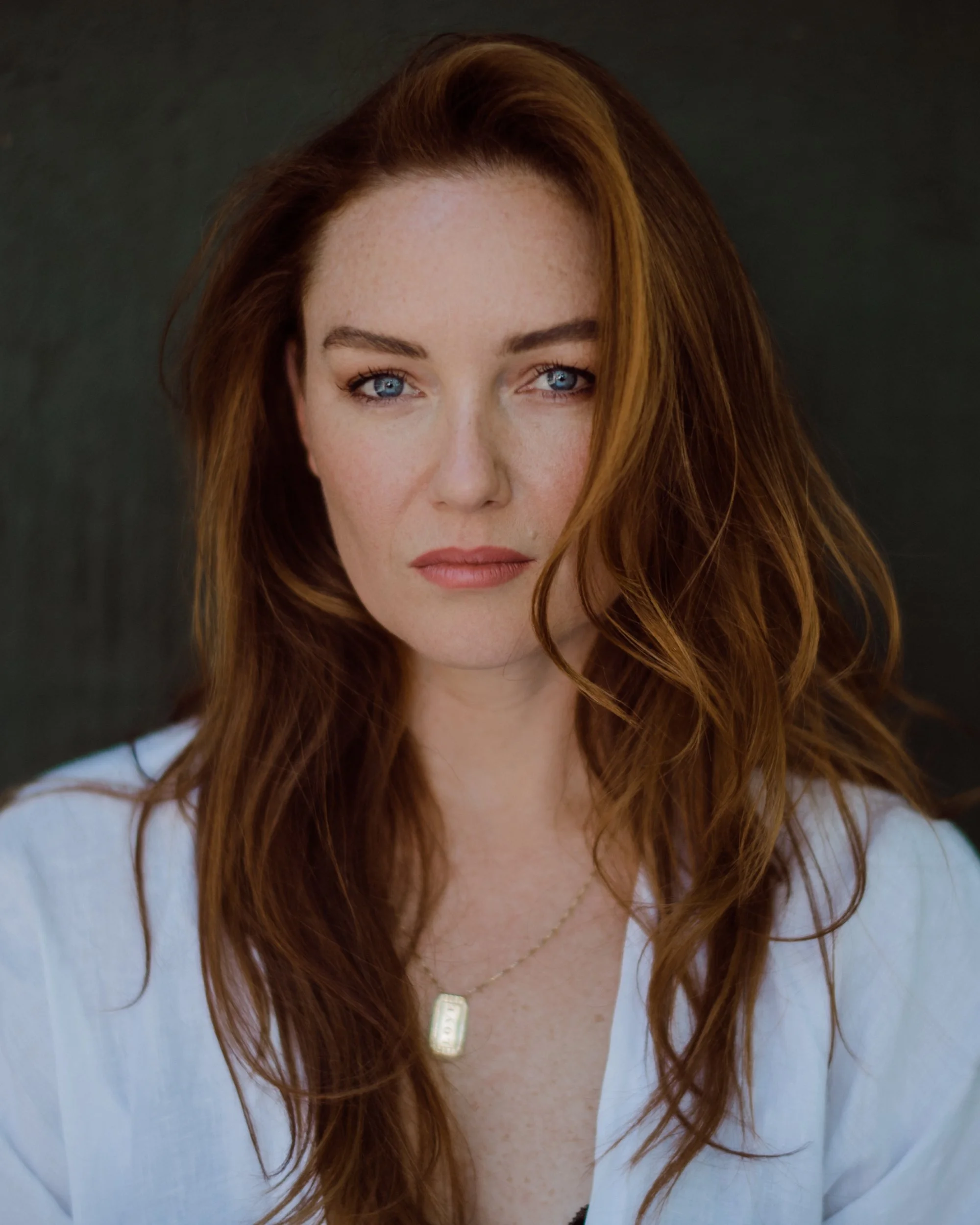 Portrait of a woman with long red hair, wearing a white top and a necklace, looking directly at the camera.
