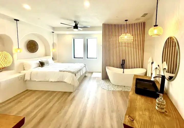 Modern bedroom with white walls, wooden flooring, and a standalone bathtub. Features include a large bed with white bedding, decorative lighting, round mirrors, and a wooden vanity with a black sink.