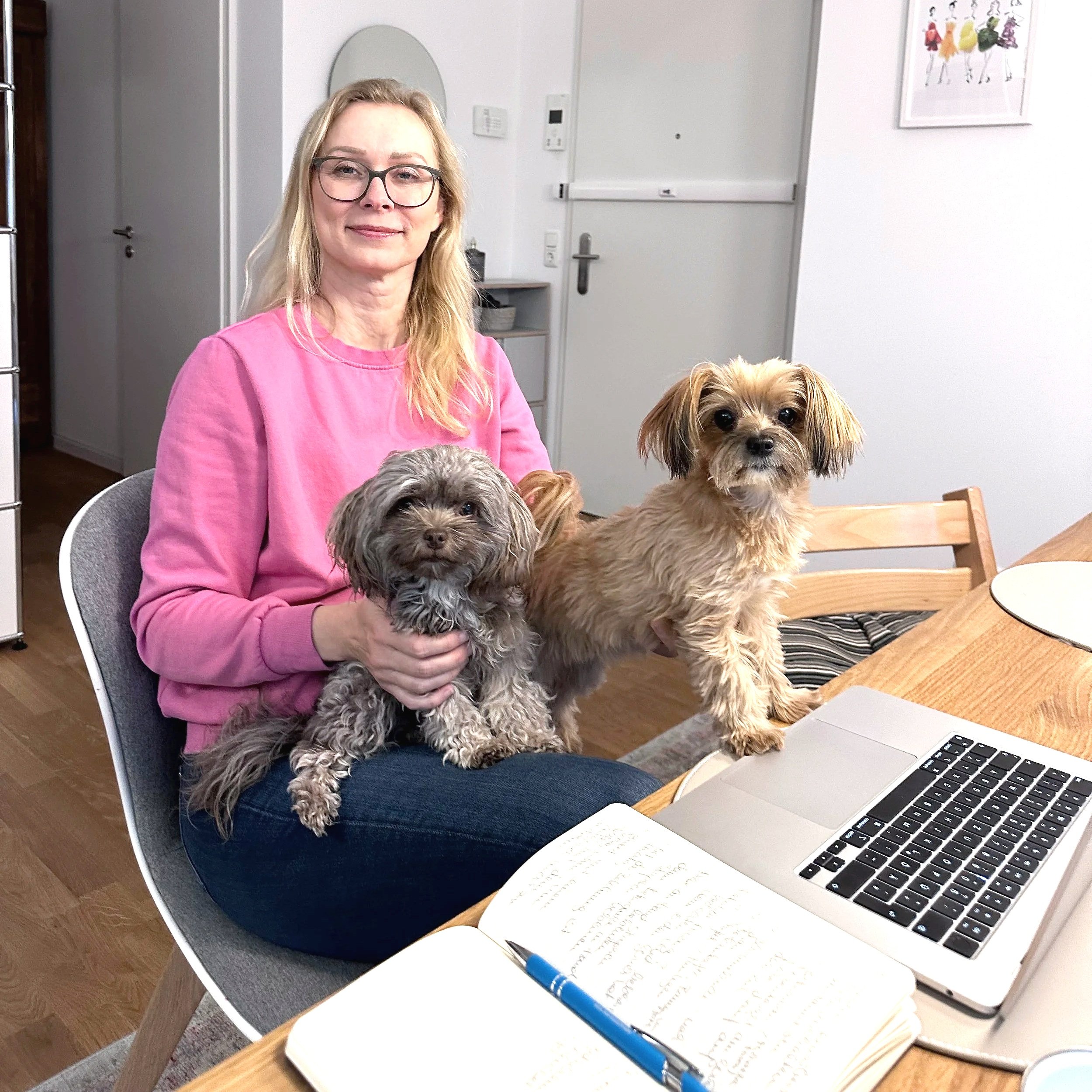 Eine blonde Frau mit Brille sitzt an einem Tisch in einer Wohnung, während sie zwei kleine Hunde hält; ein grauer, wuscheliger Hund auf ihrem Schoß und ein beige, langhaariger Hund auf dem Tisch, neben einem geöffneten Laptop und einem Notizbuch.