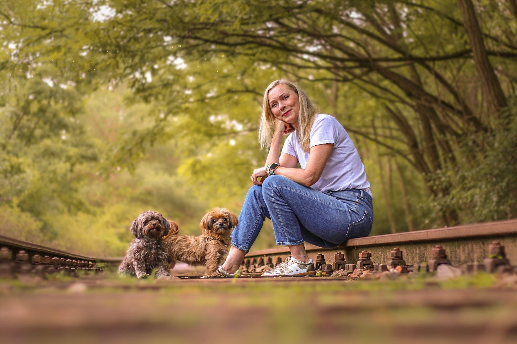 Hundetrainerin Nancy Baumeister von Dogs and the City sitzt mit zwei kleinen Hunden auf stillgelegten Bahngleisen in einem Park, umgeben von grünen Bäumen.