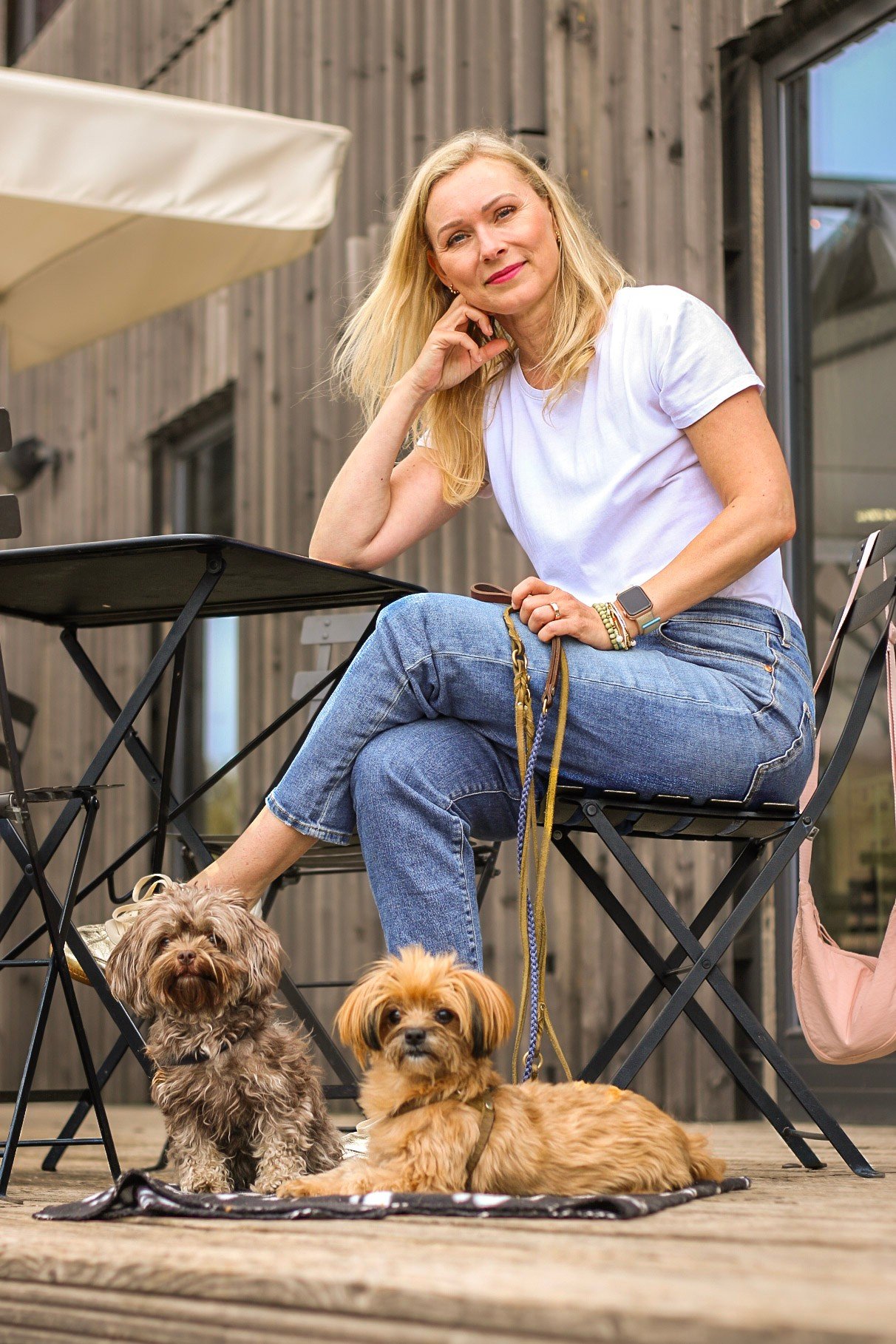 Eine blonde Frau sitzt auf einer Mikrofasertisch auf einer Terrasse mit zwei kleinen Hunden, eine braune, wellige und eine beige, kurze. Im Hintergrund ist eine Holzfassade und ein Fenster.