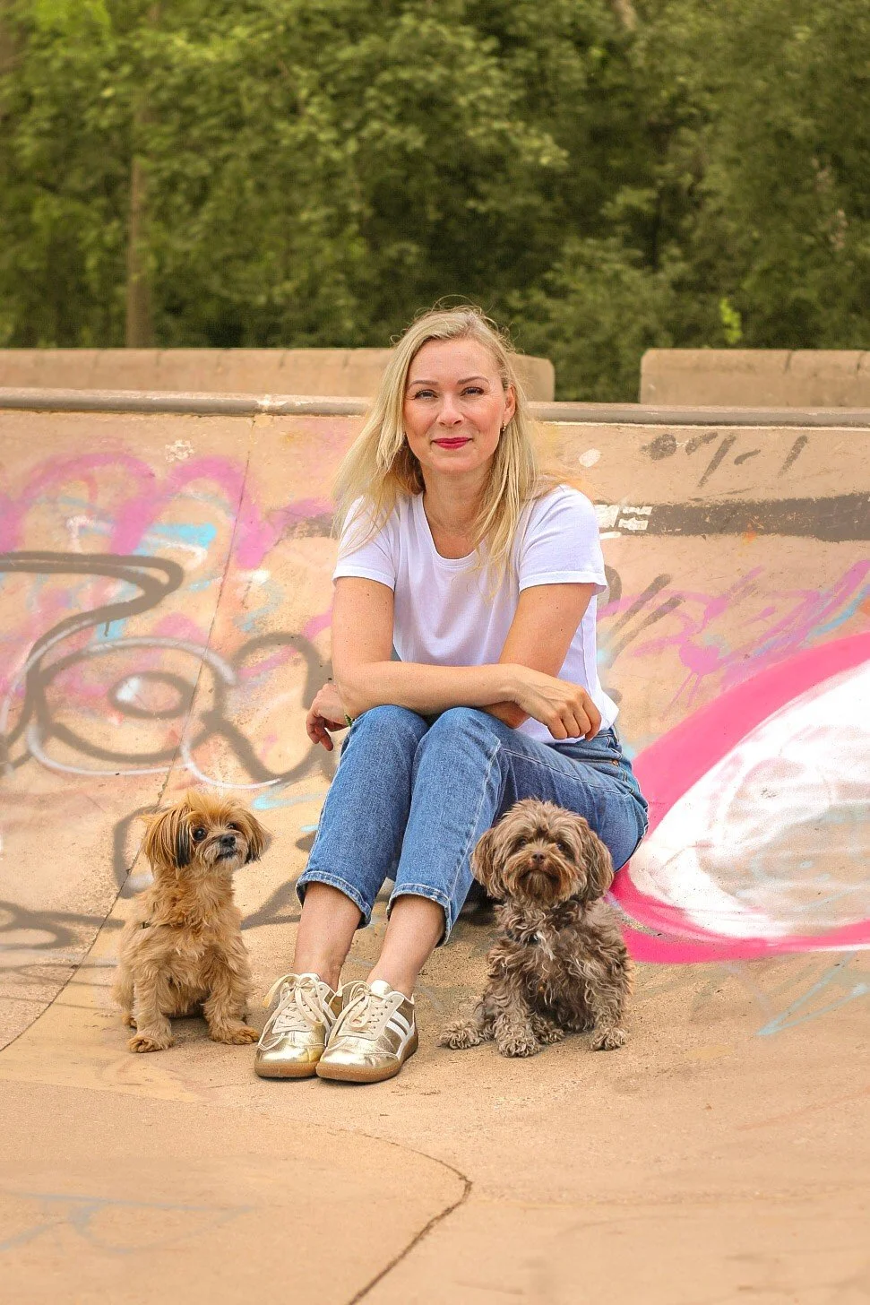 Hundetrainerin Nancy Baumeister von Dogs and the City sitzt lachend auf einer Skaterampe mit zwei kleinen Hunden, trägt ein weißes T-Shirt und Jeans, im Hintergrund grüne Bäume und Graffiti.