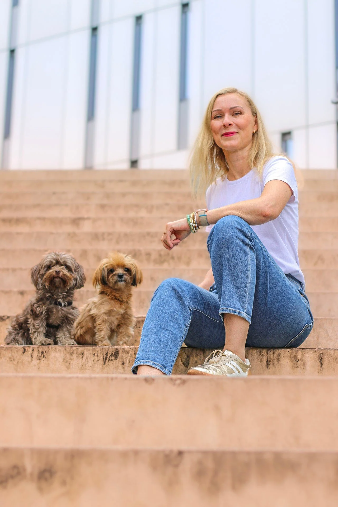 Hundetrainerin Nancy Baumeister von Dogs and the City sitzt mit zwei kleinen Hunden auf einer Treppe vor einem modernen Gebäude in Berlin.