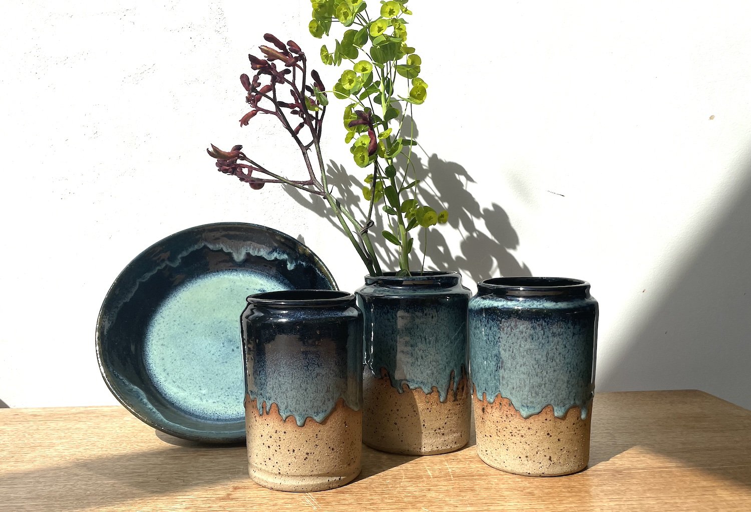 Set of three ceramic vases with floral arrangements on a wooden surface, against a white wall. The vases are glazed with blue and natural tones; one vase contains green and purple leafy stems.
