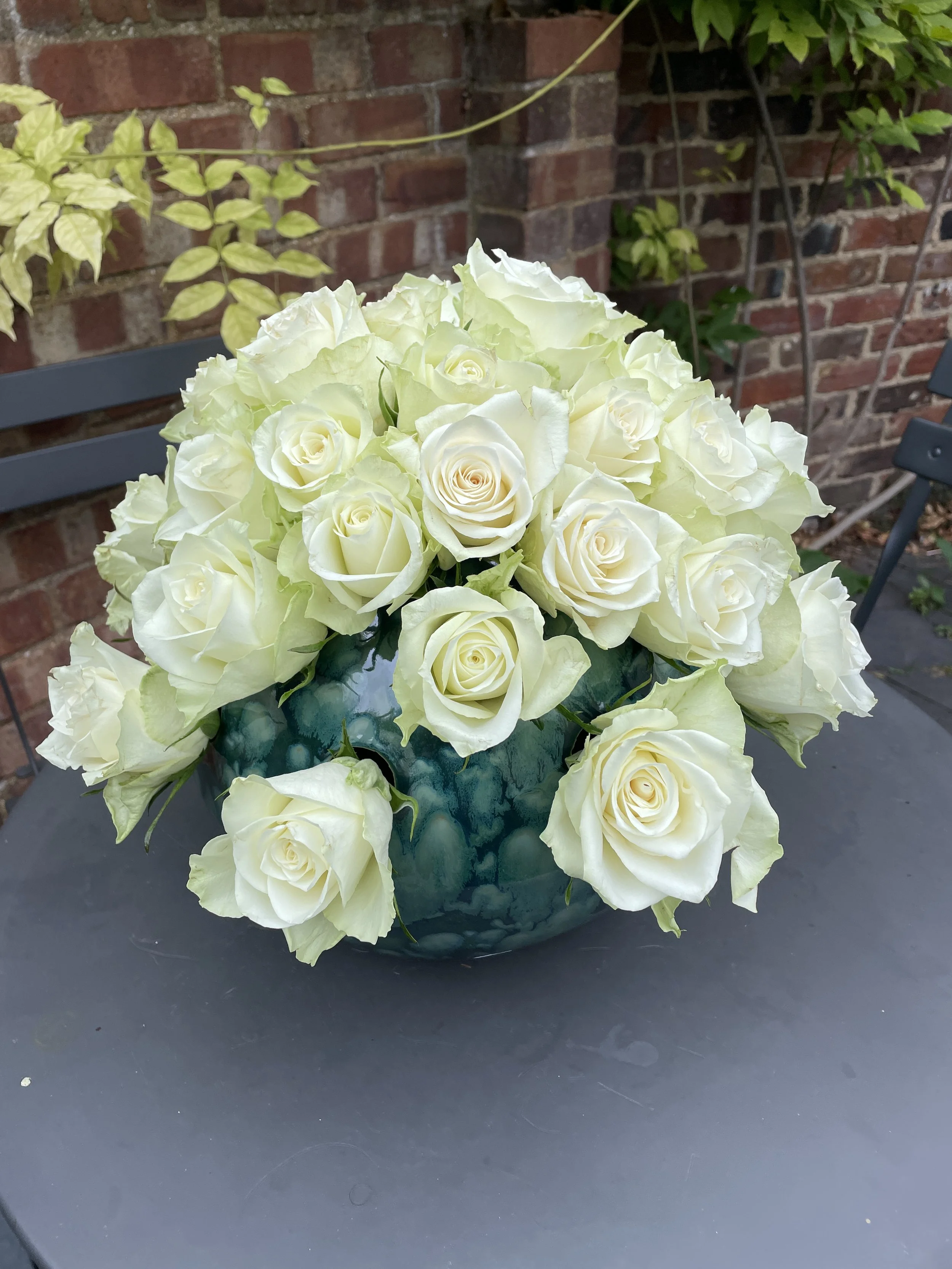 A bouquet of white roses and white flowers arranged in a dark green ceramic vase on a black table outside, with a brick wall and other greenery in the background.