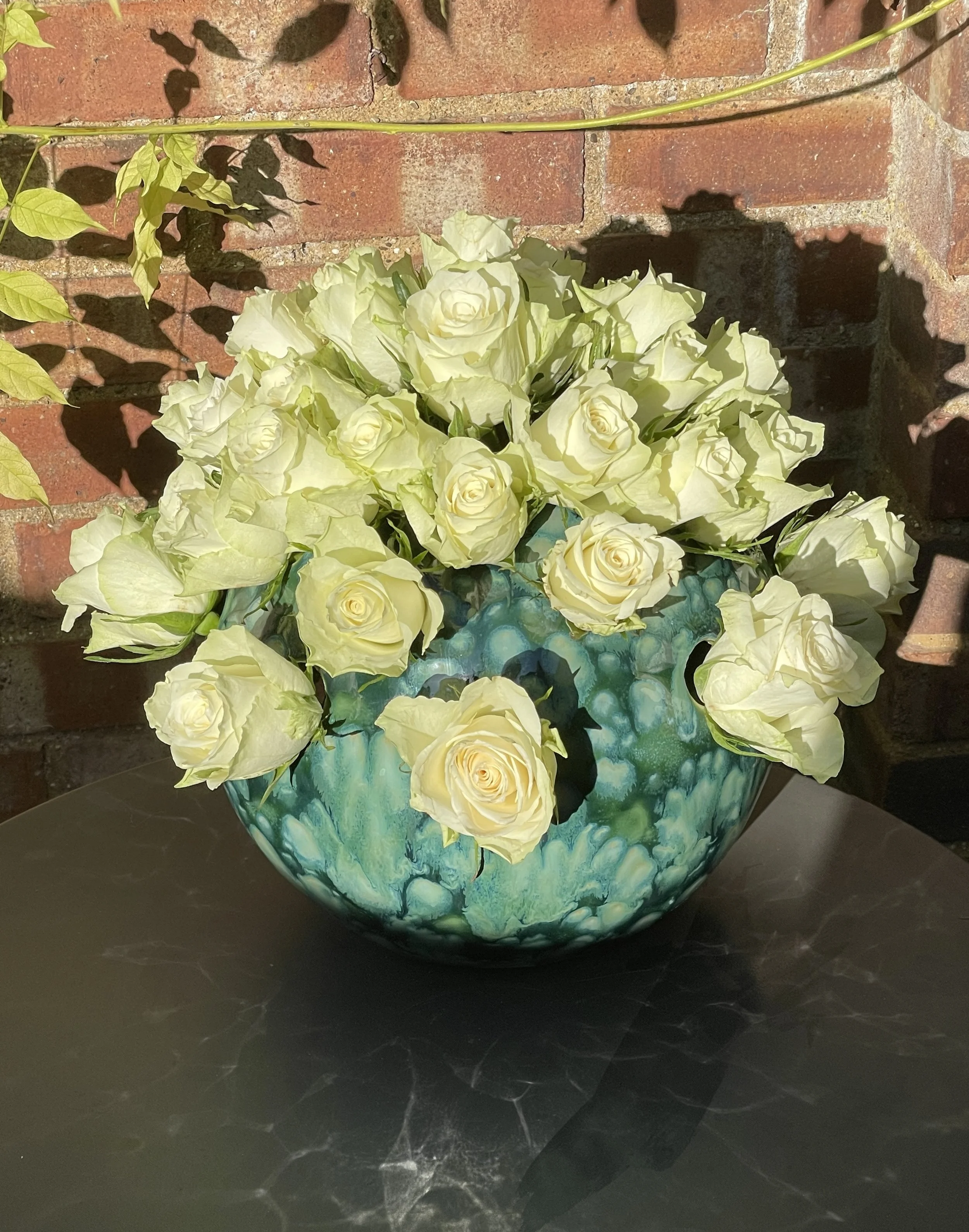 A decorative bowl filled with white roses and cream-colored flowers, placed on a dark surface against a brick wall background.