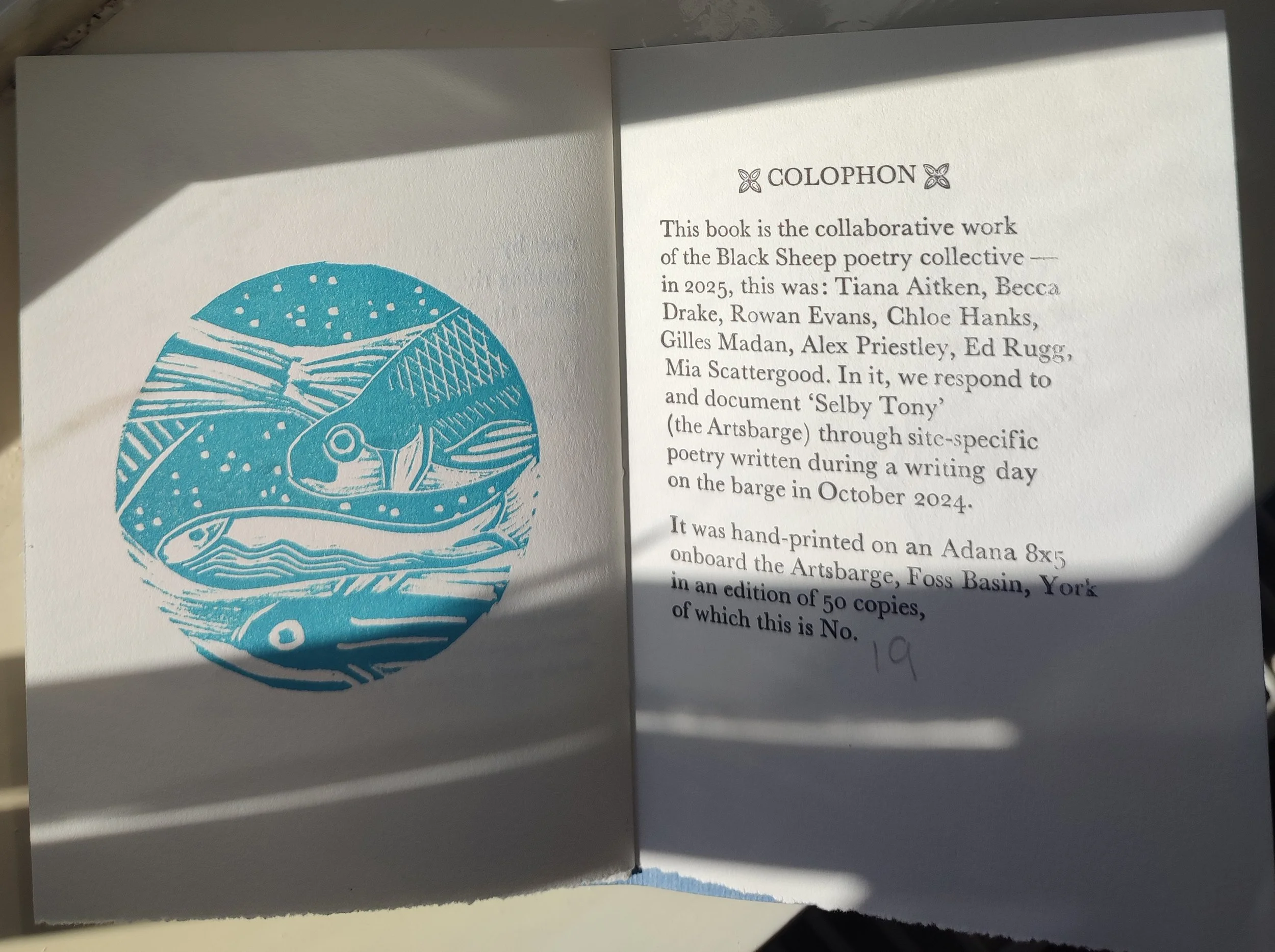 Open book with blue illustration of fish on the left page and a printed statement on the right page describing a collaborative poetry project by the Black Sheep poetry collective, with handwritten note '19' in pencil at the bottom.