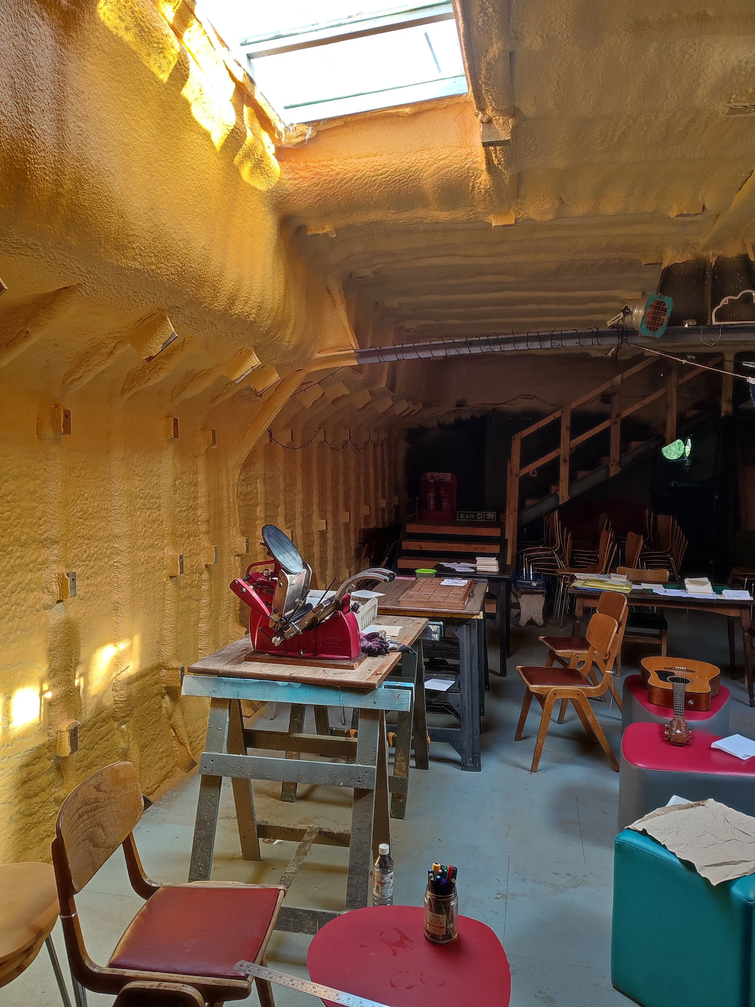Interior of a room with a yellow foam wall, work tables, chairs, a guitar, and a skylight ceiling.