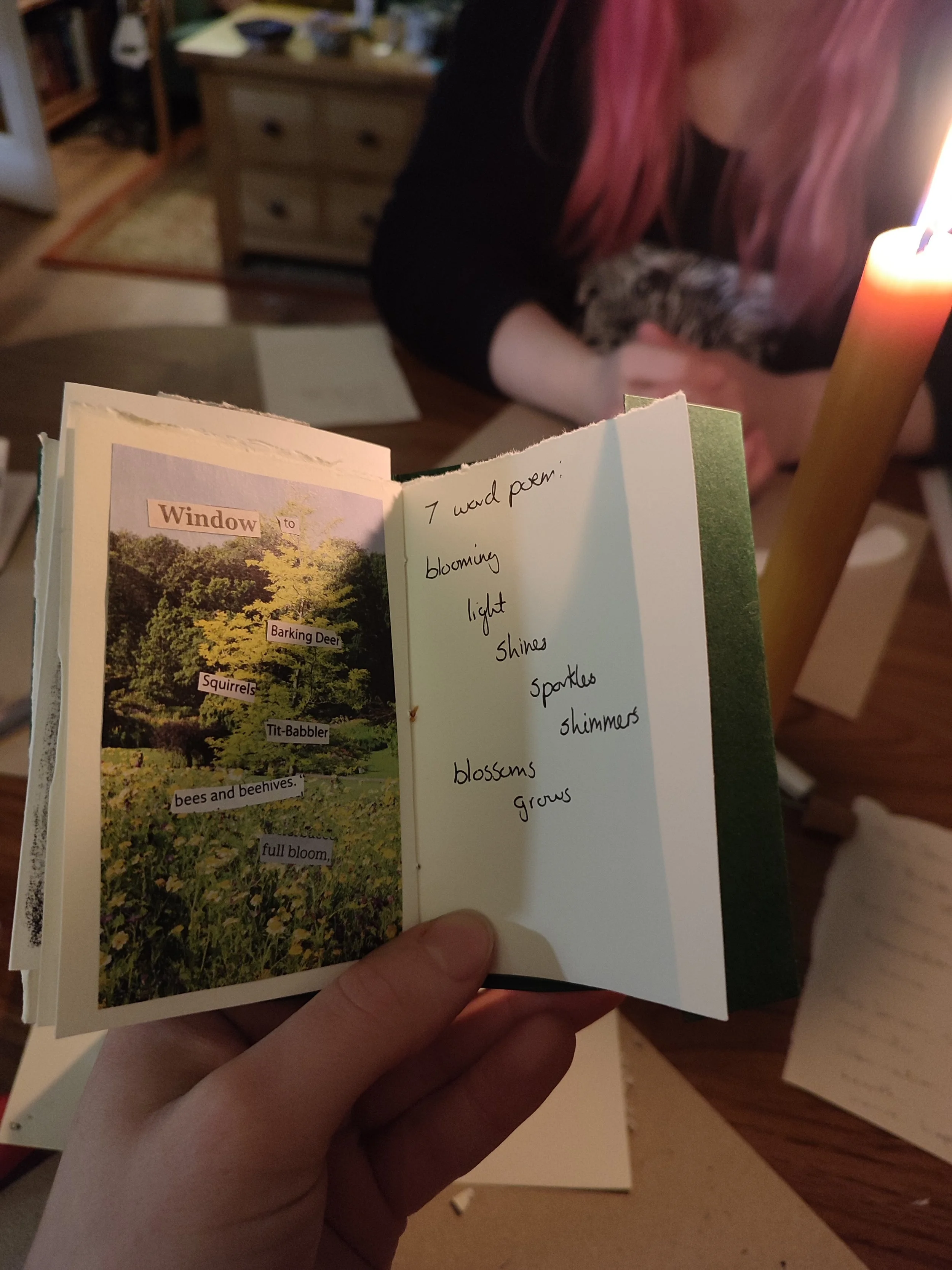 Hand holding an open handmade book with a collage on the left page showing nature images and text about a garden scene. The right page has handwritten poem lines related to blooming and growth. In the background, a person with pink hair holds a cat, and a lit candle is visible on a wooden table.
