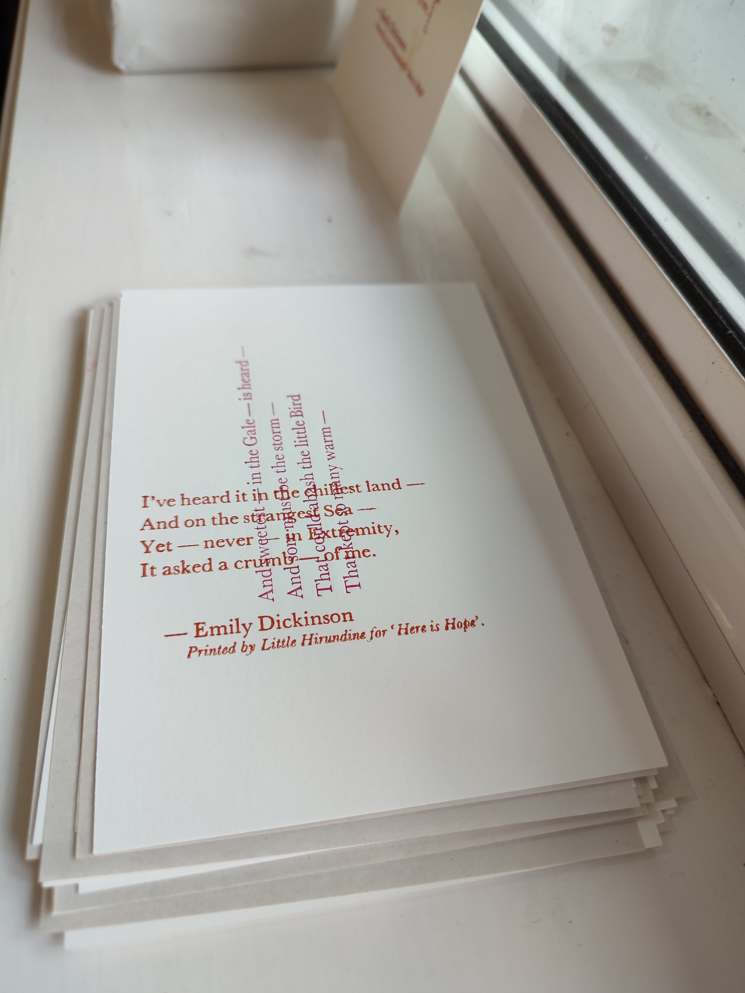 Stack of printed poems or literary pages with a poem by Emily Dickinson, placed on a white surface near a window.