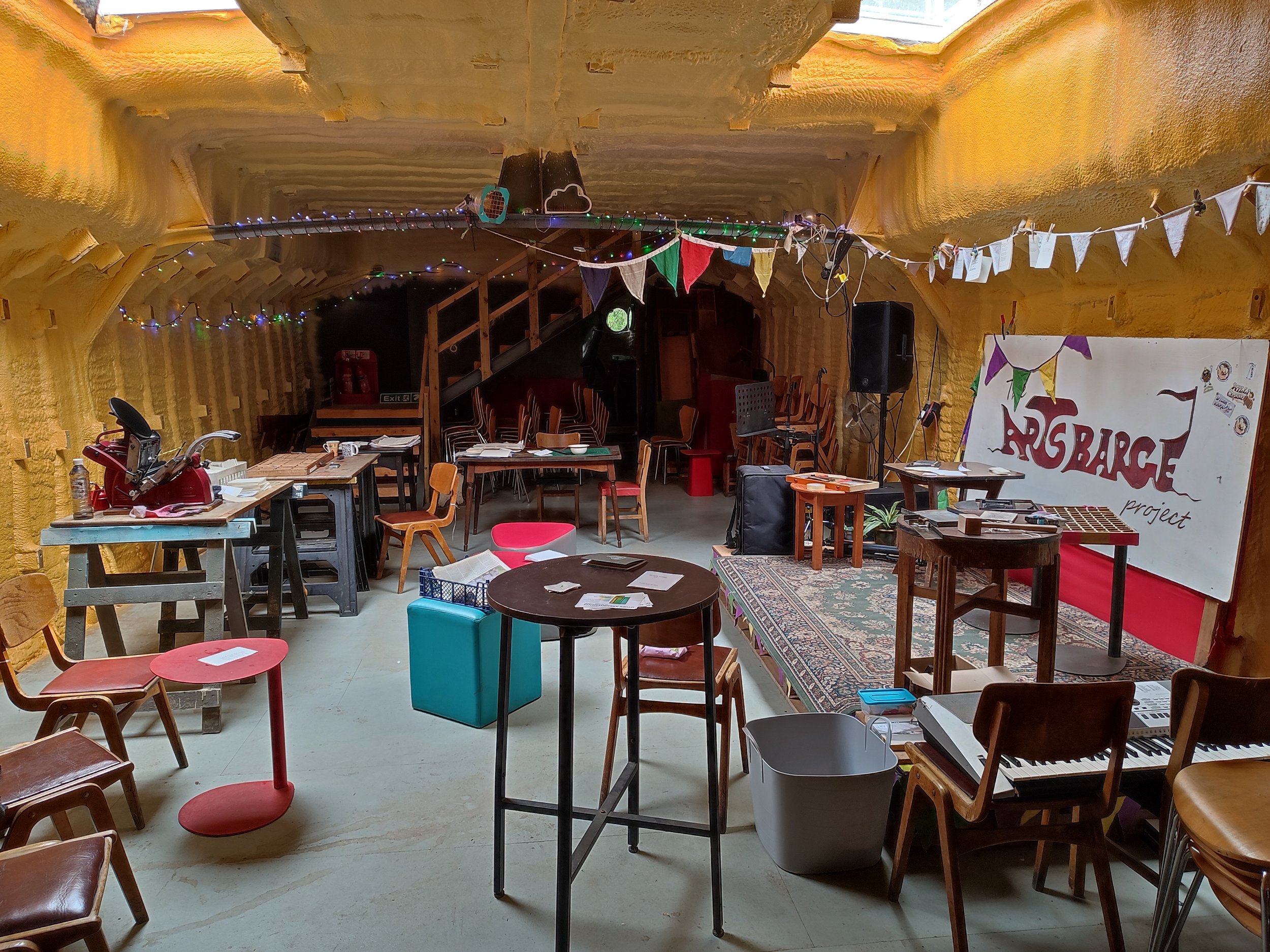 An indoor space with yellow walls, decorated with colorful bunting and string lights. There are various tables and chairs, a keyboard, and a large sign that reads "MAGBARAGE project." The space appears to be set up for a gathering or event.
