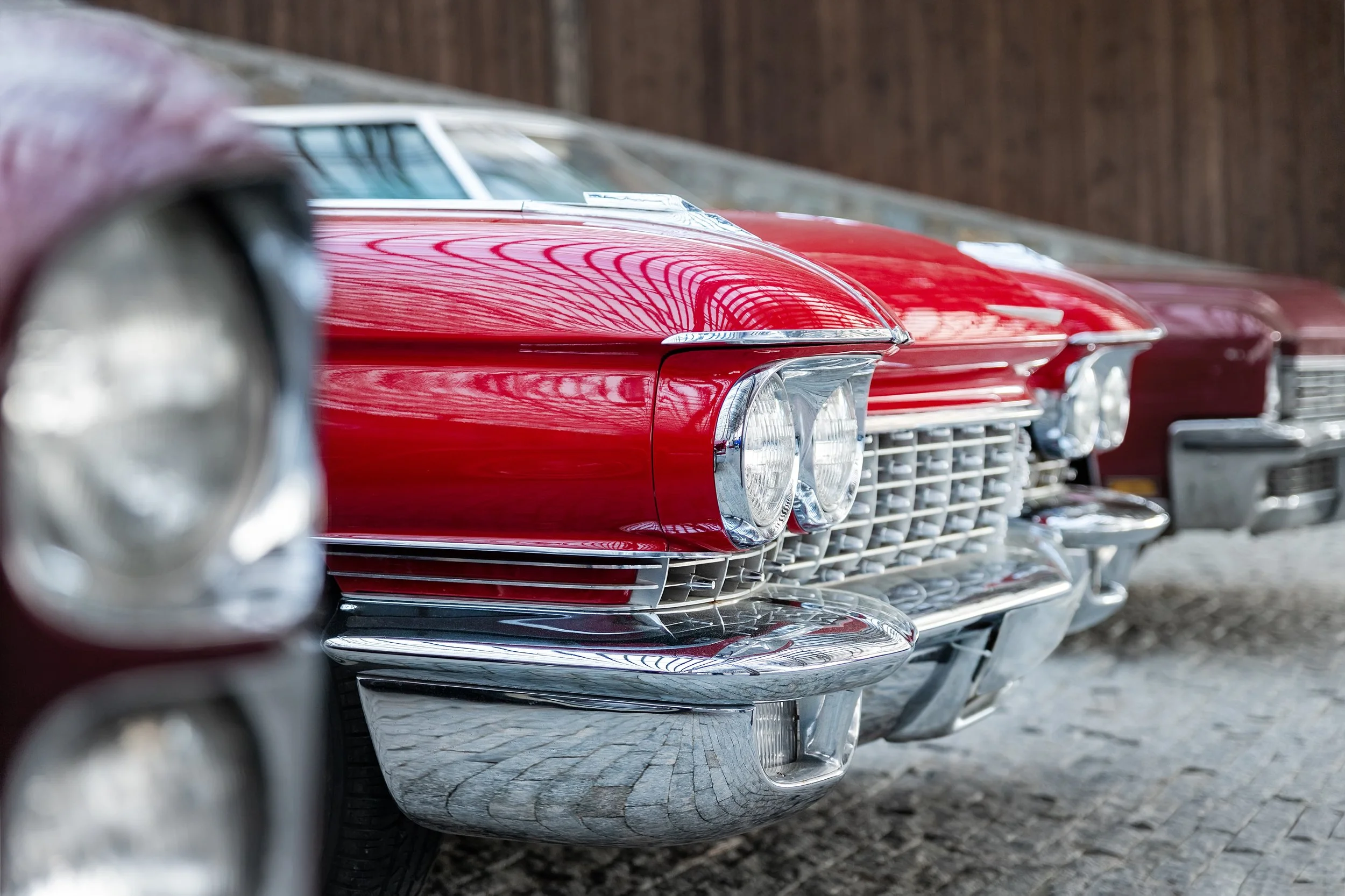 Close-up van de voorzijde van meerdere klassieke rode en zilveren auto's met chroom details, geparkeerd naast een houten muur.