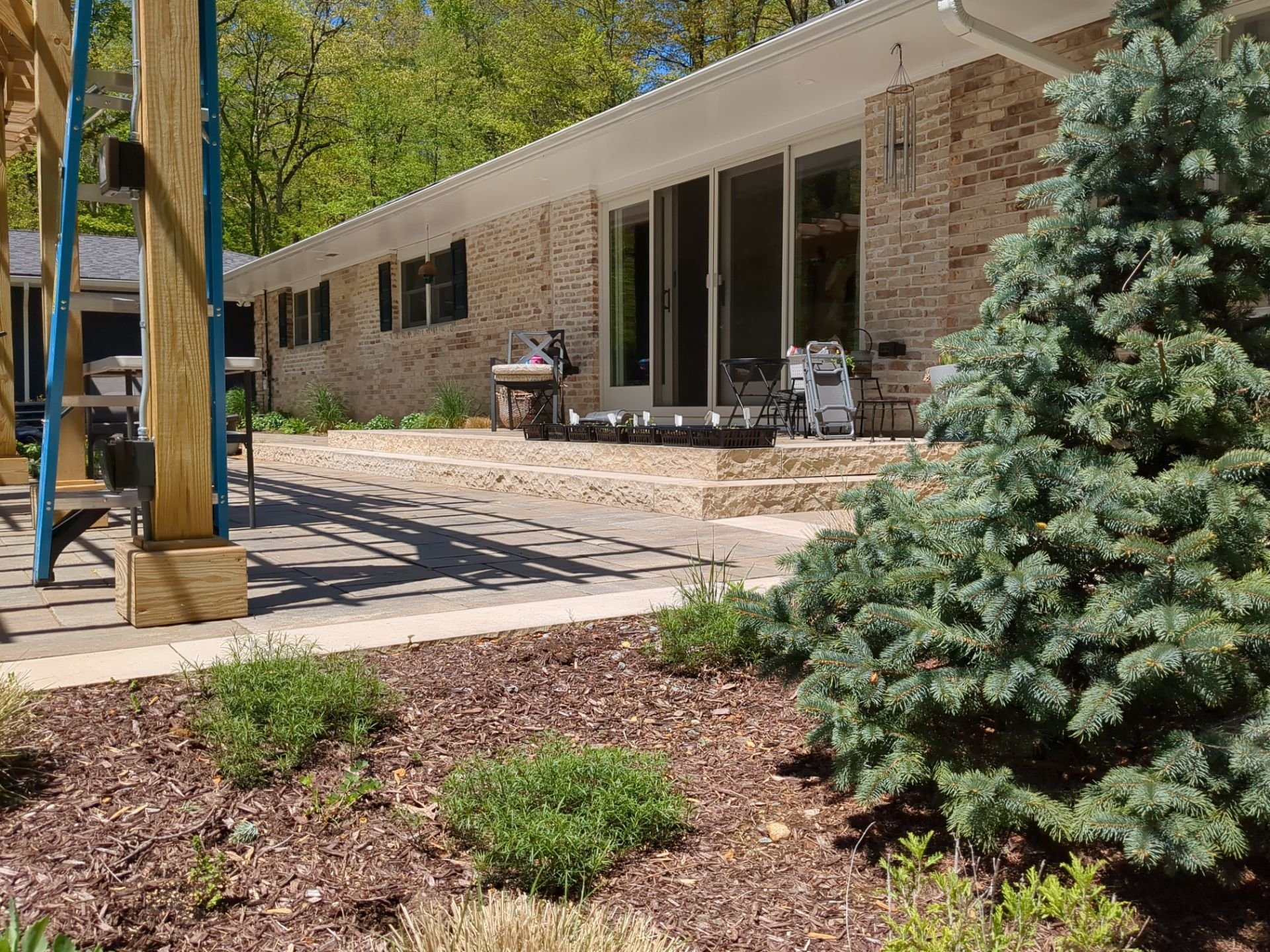 Backyard landscape with evergreen tree, perennial garden beds, stone patio, and pergola beside the home