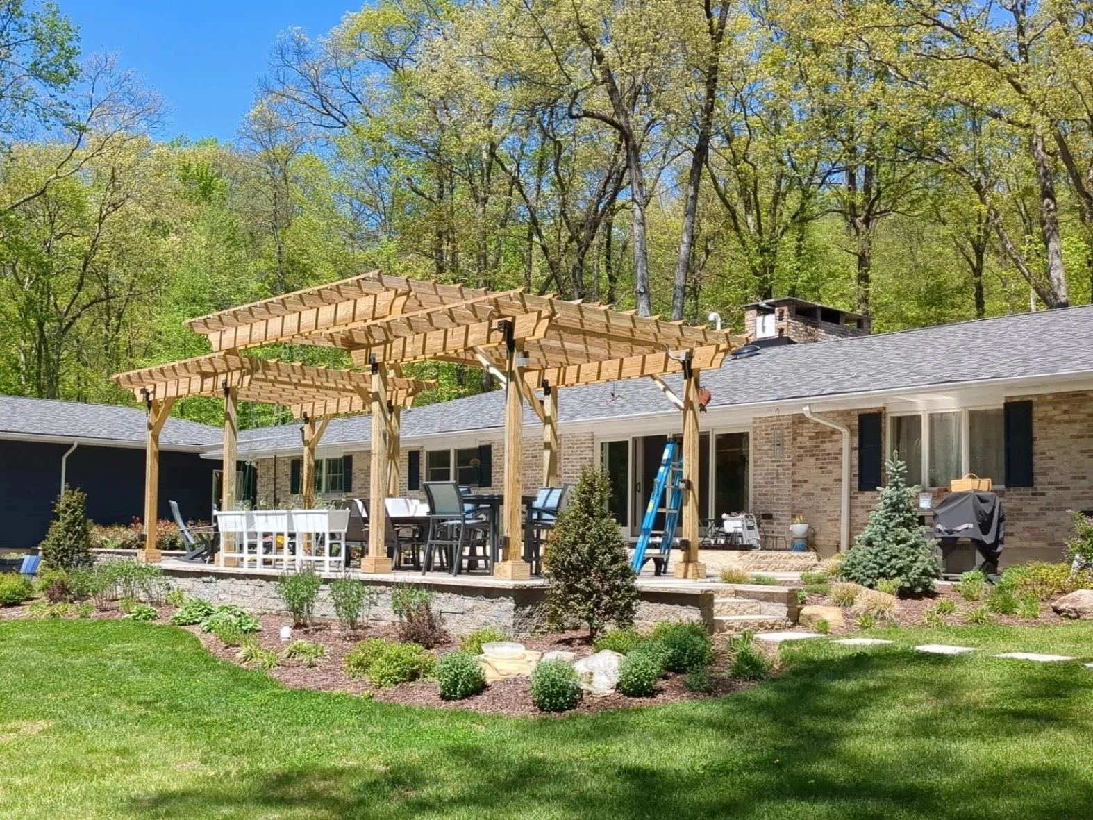 Landscaped backyard with pergola, stone terrace, layered garden beds, and lawn surrounding a renovated home