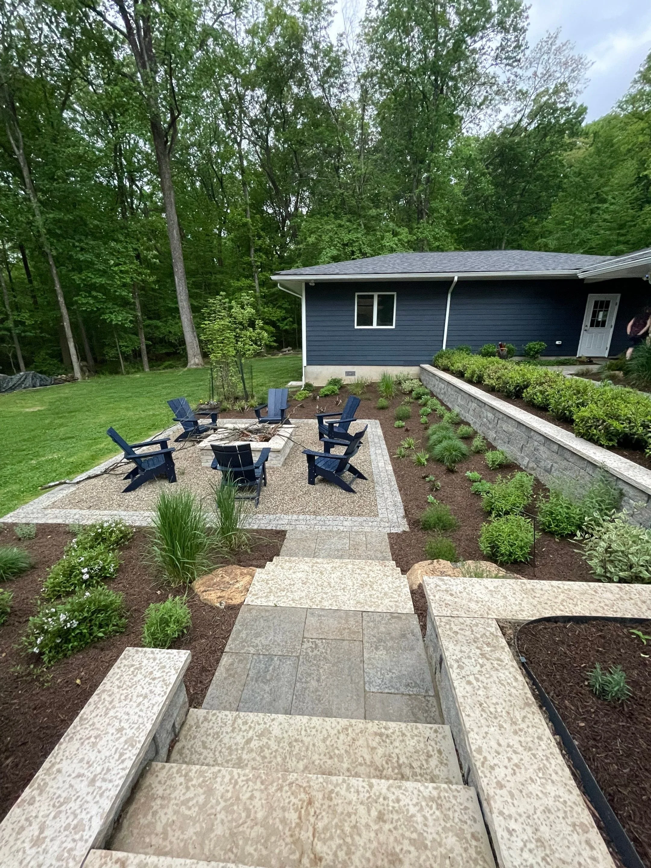 Backyard fire pit patio surrounded by pea gravel, Adirondack chairs, stone pathways, and layered garden beds