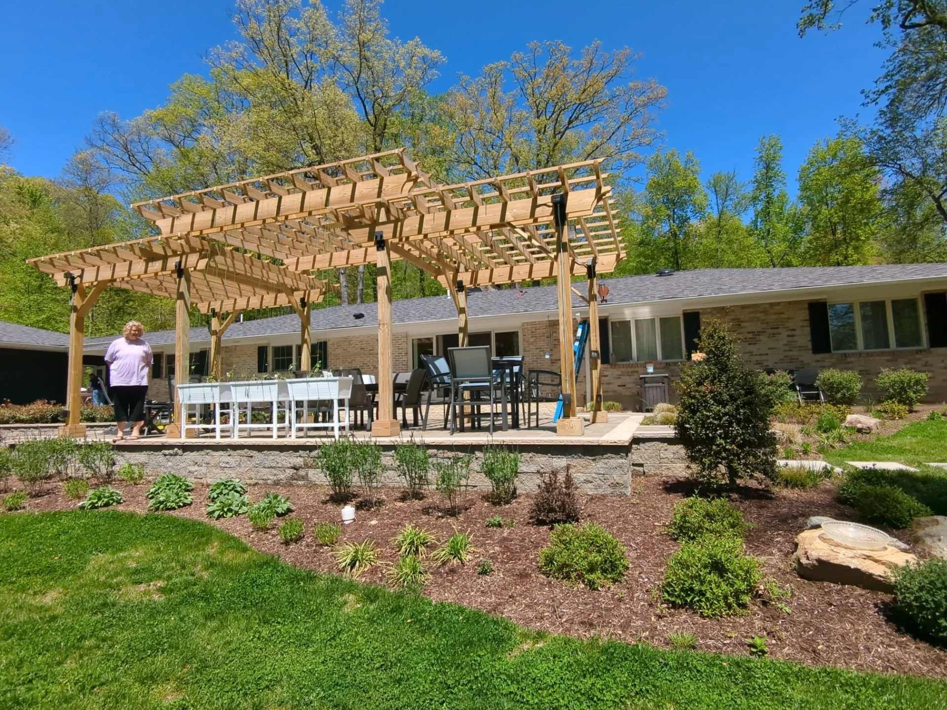 Backyard landscape with wooden pergola, stone patio, layered perennial gardens, and lawn surrounding the home