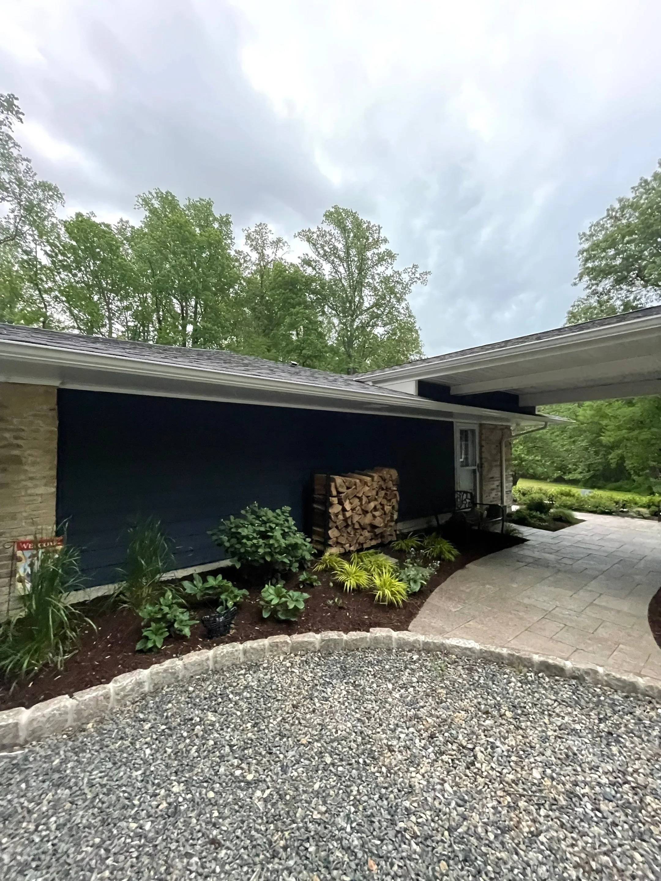 Garage walkway with stacked firewood, landscaped garden beds, ornamental grasses, and stone paver path