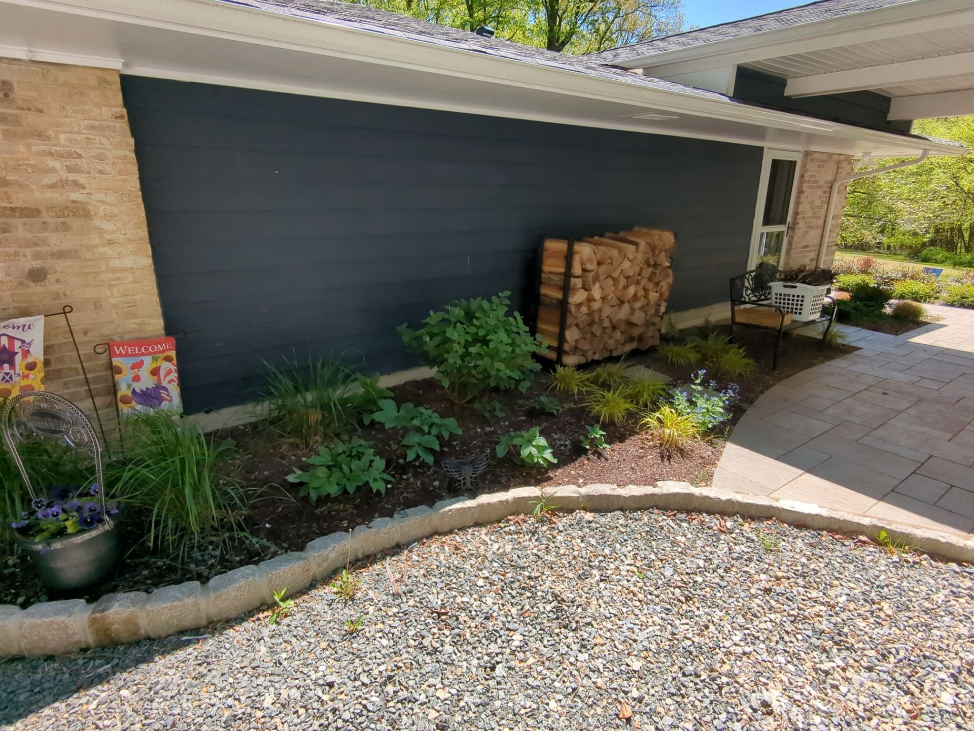 Side yard with stone paver walkway, curved garden bed, ornamental plantings, and stacked firewood
