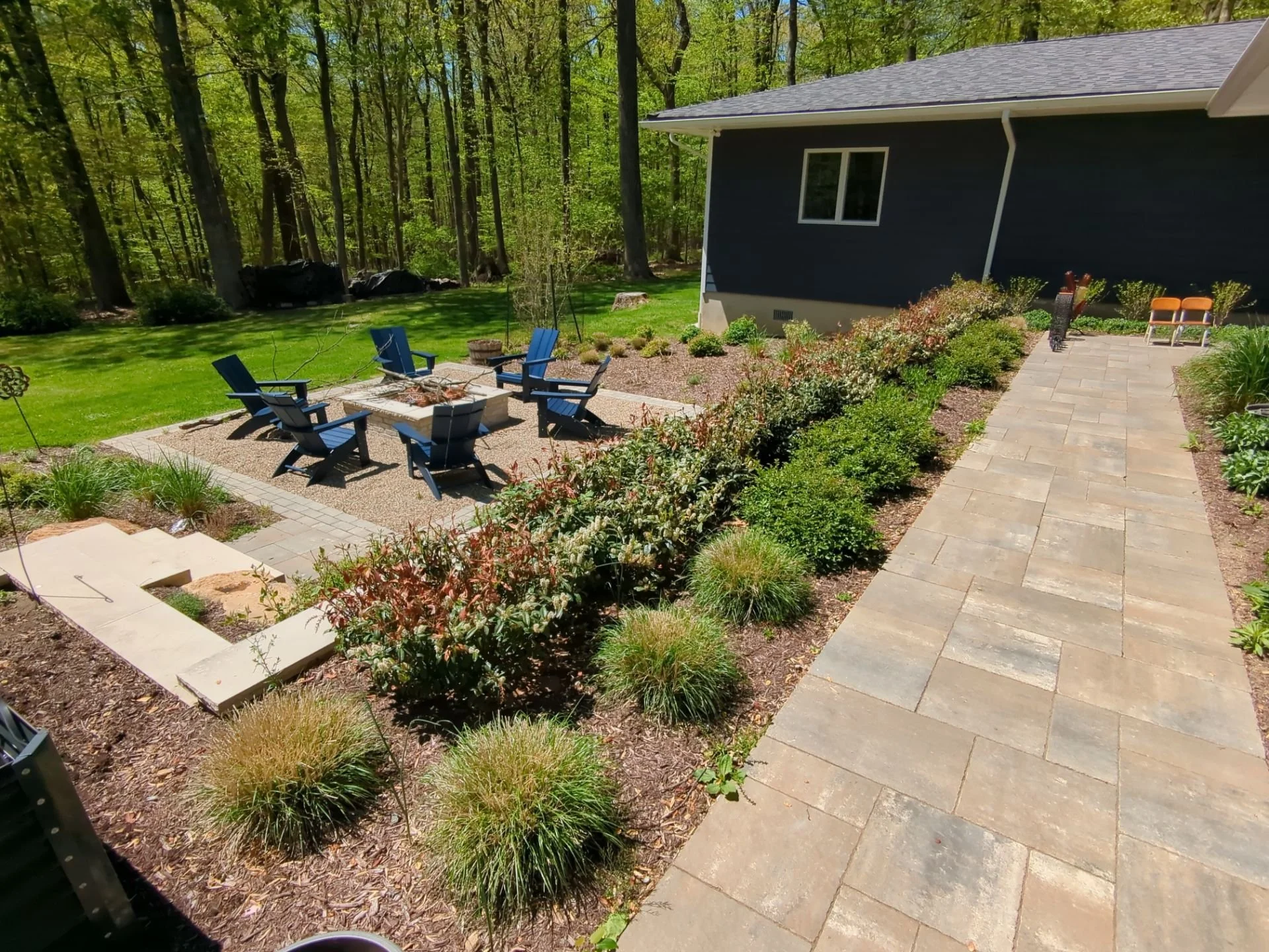Backyard landscape with stone walkway, fire pit patio, layered garden beds, and outdoor seating surrounded by trees
