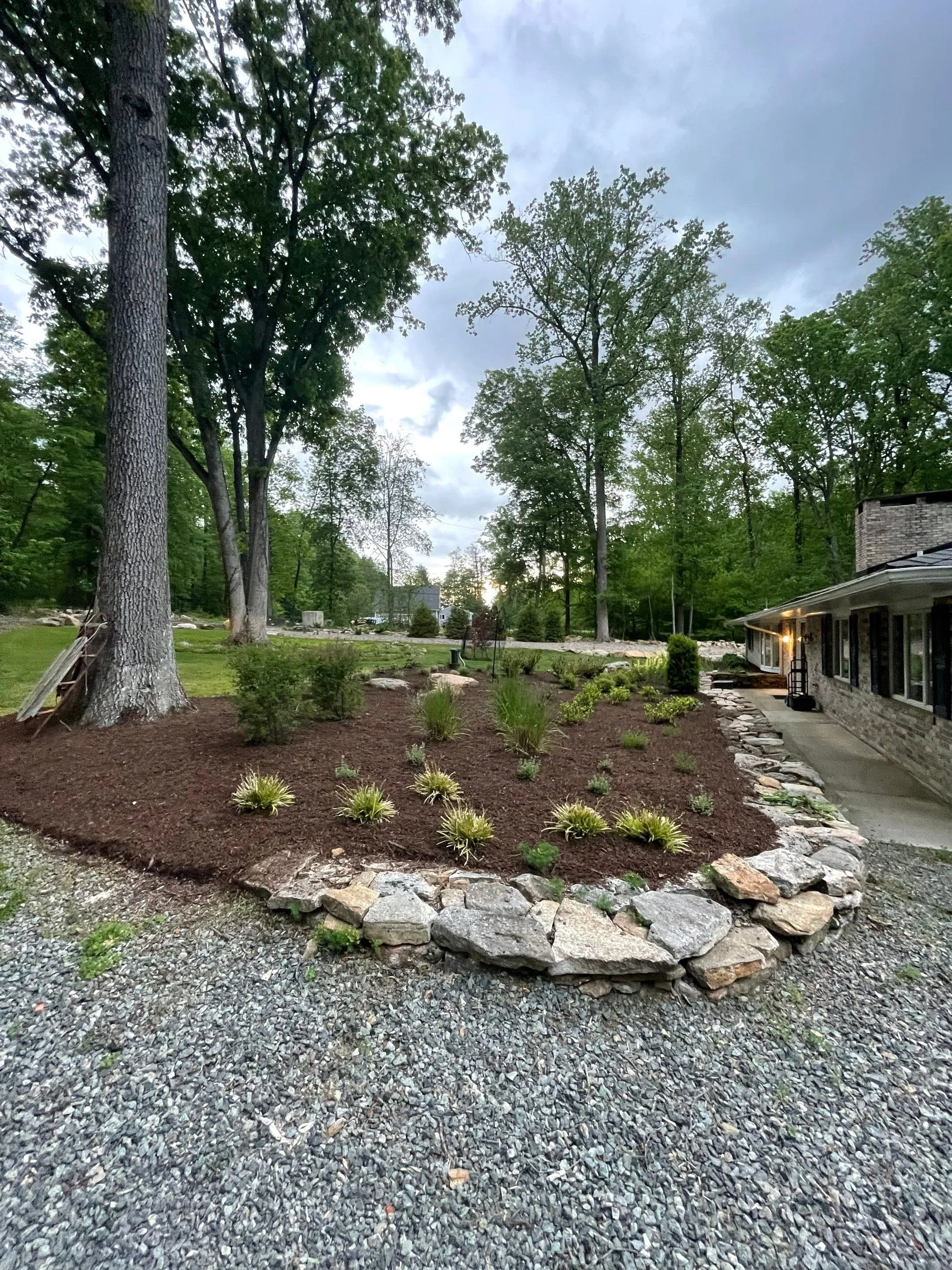 Front landscape with mature trees, natural stone edging, layered plantings, and a curved garden bed