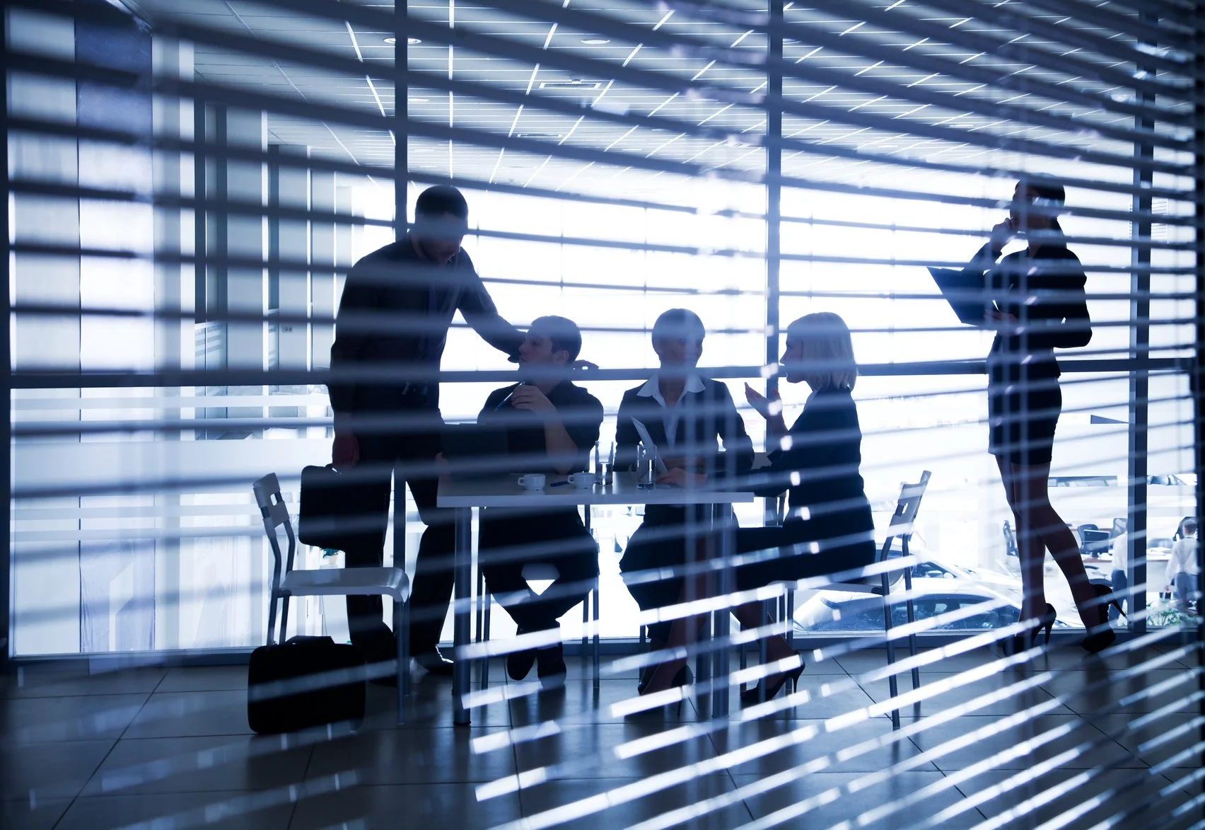 Silhouettes of business professionals in a modern office seen through window blinds, with some standing and talking, and others sitting at a table, with one person on the phone.