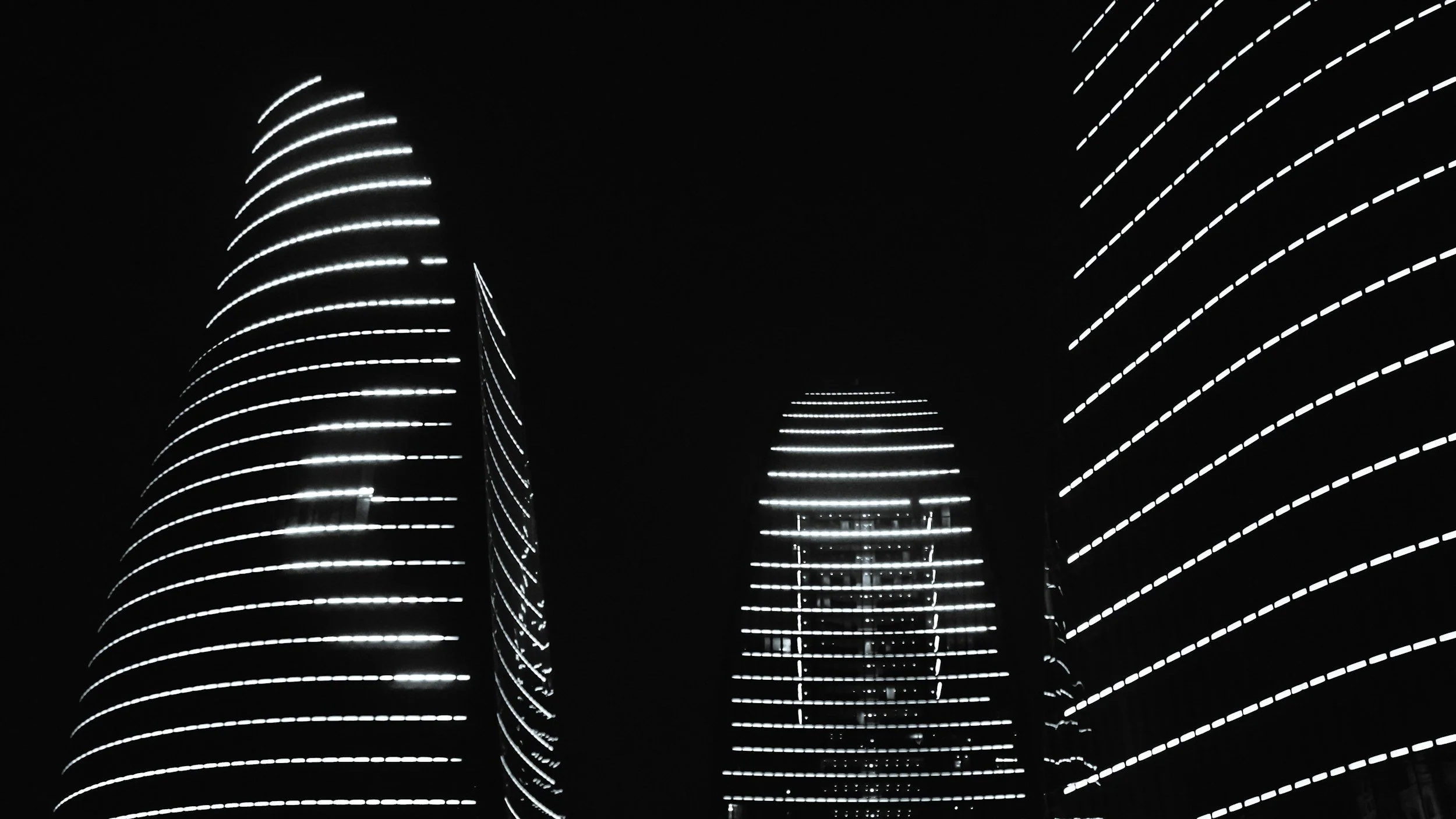 Night view of three modern skyscrapers with illuminated horizontal white lines spiraling upwards on dark background.