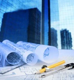 Architectural blueprints and rolled-up plans on a desk in front of a window with city buildings outside.