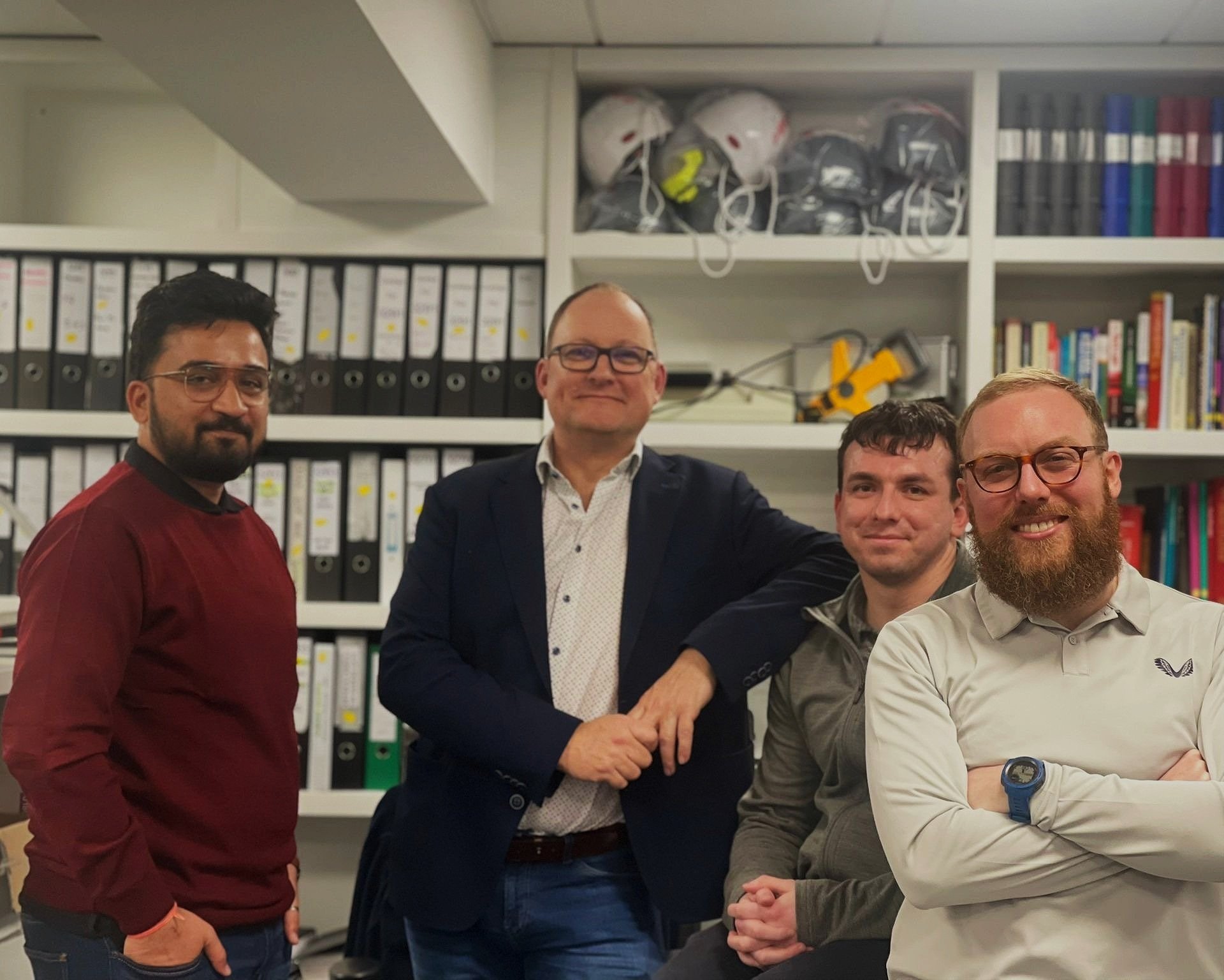 Four men standing in an office, two shaking hands, with shelves of binders, books, and helmets in the background.
