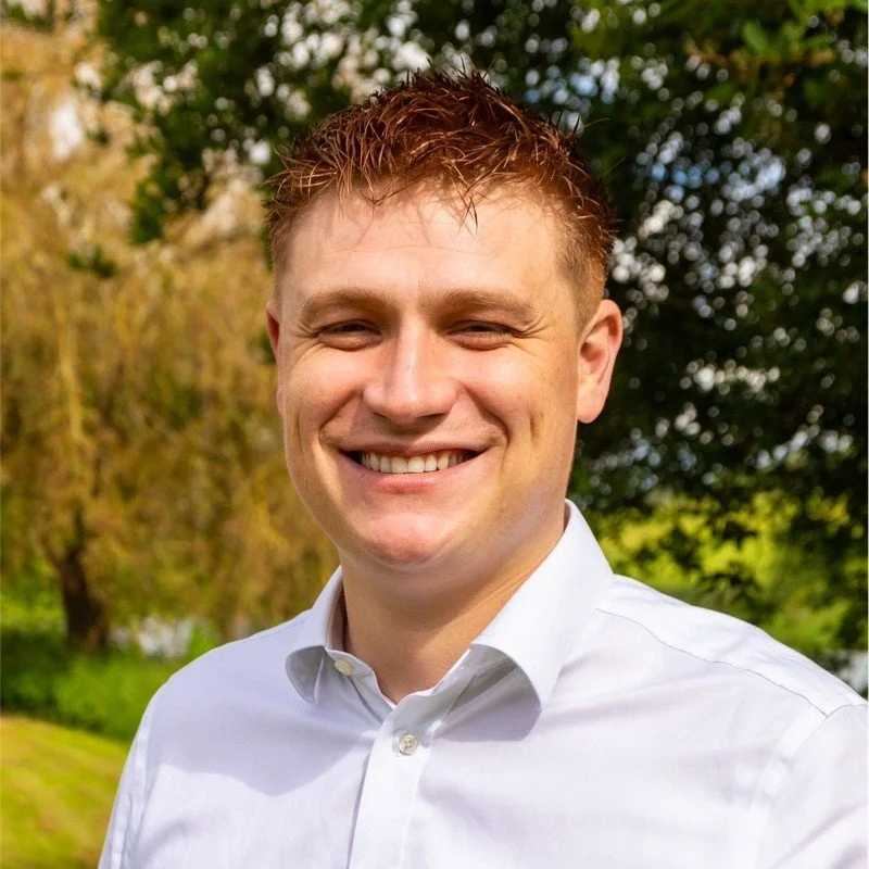Young man smiling outdoors with trees and greenery in the background
