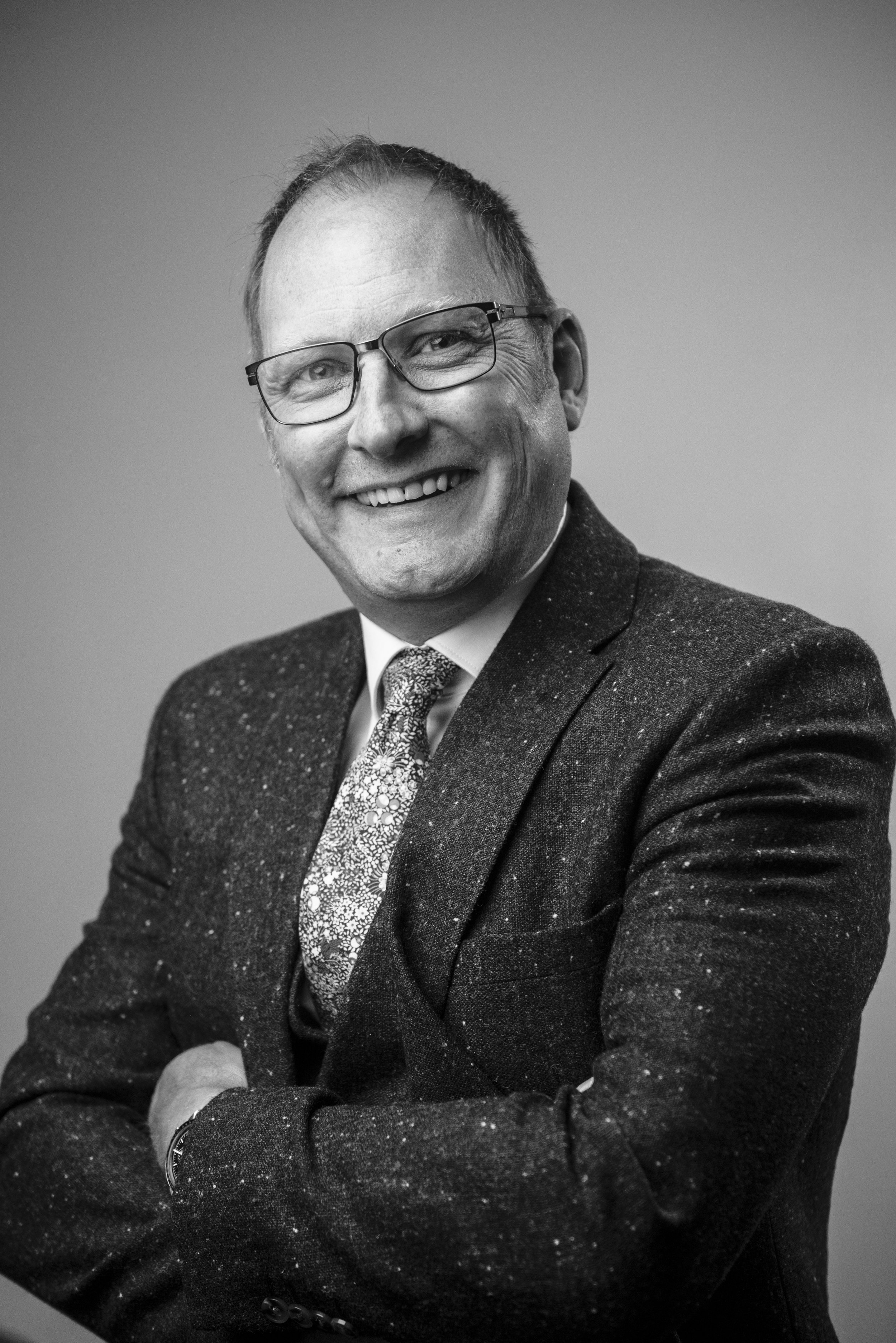 Black and white portrait of a smiling man wearing glasses, a patterned tie, and a dark blazer with speckles.