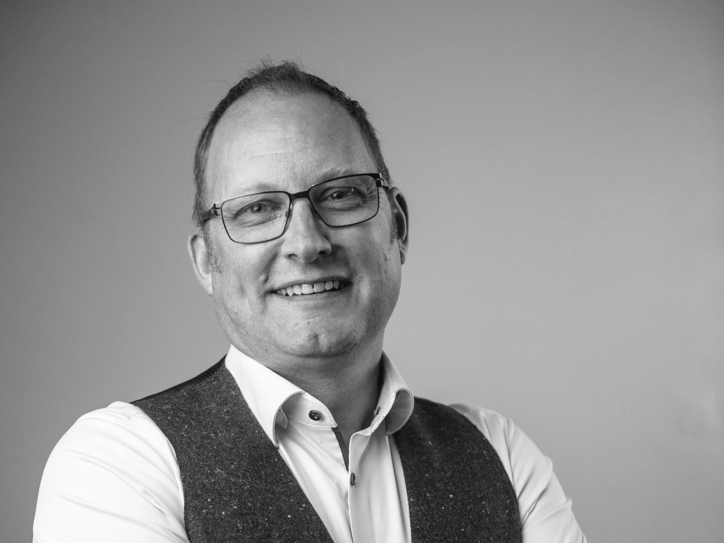 Black-and-white portrait of a smiling man wearing glasses, a white shirt, and a dark vest, with arms crossed against a plain background.