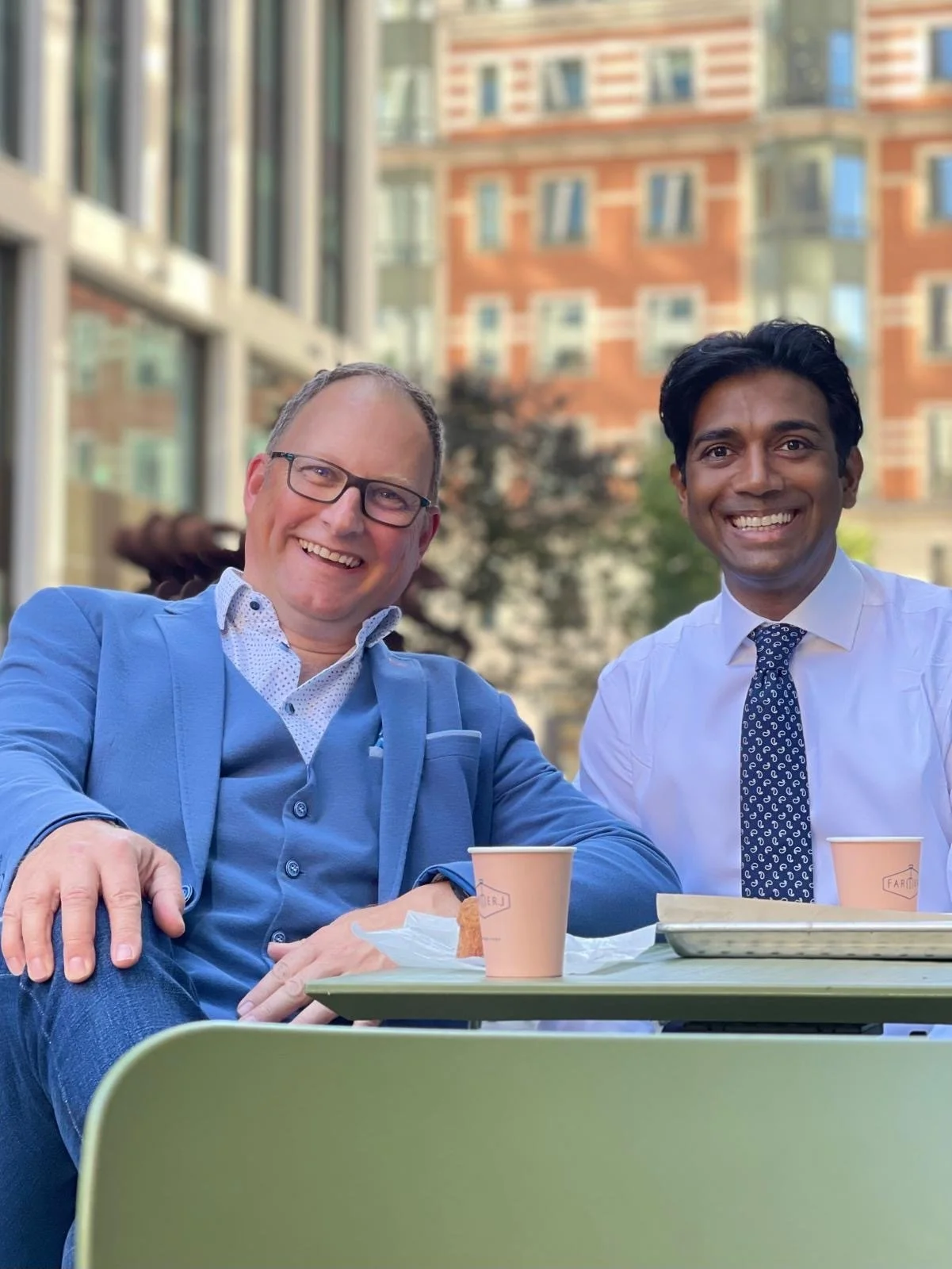 Two smiling businessmen, one in a blue suit and glasses, and the other in a white shirt with a patterned tie, sitting at an outdoor table with coffee cups and food, with a cityscape of buildings in the background.